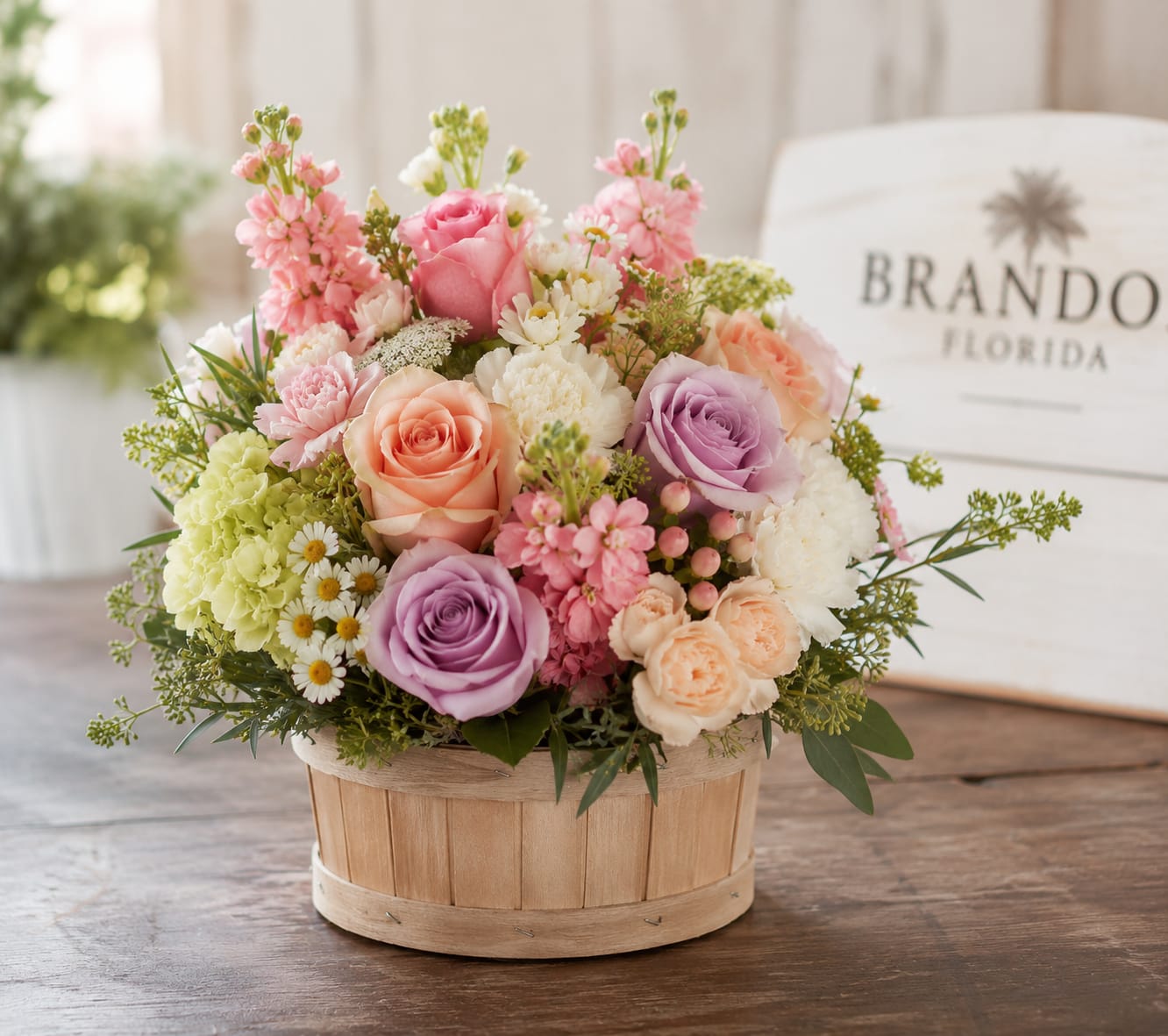 Basket of Beauties for Brandon  - Pastel roses take center stage in this compact, rounded flower arrangement-perfect as a birthday bouquet, Mother's Day gift, or a sweet anniversary surprise. The Basket of Beauties for Brandon reads as a romantic, cheerful gift that will make the person you're thinking of feel genuinely seen: soft pinks and peaches layered with lavender and white create a delicate, uplifting impression that says celebration and care.  Roses are paired with ruffled carnations and small daisy-like filler blooms, giving the design a garden-style texture and an airy, approachable silhouette. Pale green accents add fresh contrast while the handcrafted wooden basket lends warm, rustic charm and a bit of casual elegance to tabletops or entryway surfaces. The arrangement is compact and rounded-easy to display anywhere that needs a gentle pop of spring color.  Ideal for spring occasions, birthdays, anniversaries, or simply to brighten someone's day, this basket sends a thoughtful, timeless message. Care is straightforward: keep blooms in a cool spot away from direct heat, refresh the water as needed, and enjoy the soft fragrance and layered textures for several days.