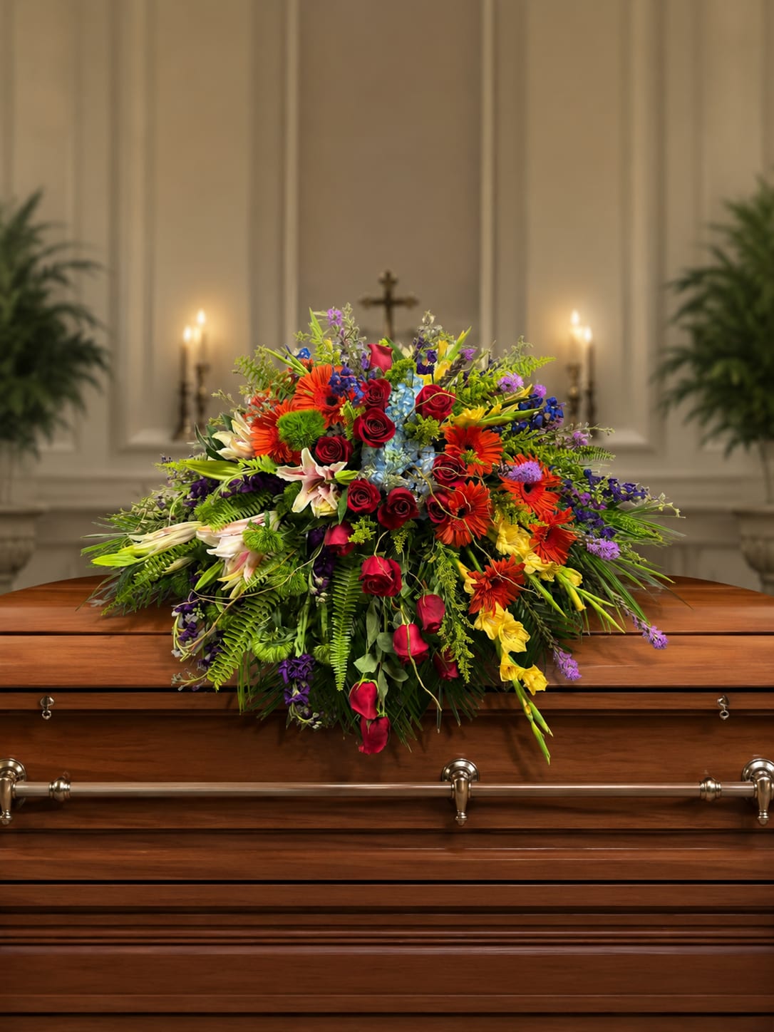 Renoir’s Touch of Vibrancy Casket Piece by Petal Perfection Flower Company  - A low, elongated spray awash in a rainbow of blooms-this funeral casket piece and sympathy arrangement brings a lively, uplifting presence to a memorial or celebration of life. The horizontal oval shape and slightly domed profile make it ideal for placement on a casket or a memorial table, while the bright palette of red, orange, yellow, blue, purple, pink, peach, lavender and green reads as a joyful tribute for someone remembered with warmth.  Closely clustered red roses, orange gerbera daisies, pink lilies, blue hydrangea and delphinium form a dense, layered composition with accents of chrysanthemums, stock and mixed seasonal flowers for depth and texture. The mix of large focal flowers and softer filler blooms creates visual movement across the arrangement, and its compact, low silhouette ensures the piece sits gracefully without obscuring views during a service.  Appropriate year-round but especially fitting for a colorful sympathy or celebration-of-life service, this arrangement also translates to bright birthday or get-well displays for someone who loved bold color. Sizes and exact flower counts vary by availability; our florists arrange and secure each spray for stable transport and display-please contact us for specific sizing or personalization requests.