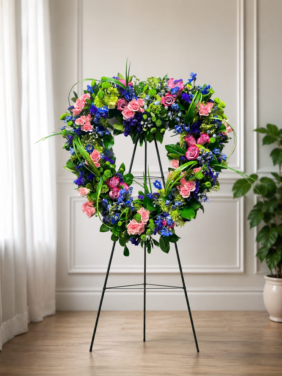 My Heart Goes Out Standing Heart - A heart-shaped standing spray that speaks of remembrance and gentle love, perfect as a sympathy or funeral tribute. The heart silhouette and open center create a tender focal point that lets the person you're honoring feel cherished and remembered, while the mix of colors-from soft pinks to vivid blues-gives the design a comforting, hopeful presence.  Clustered spray roses form the main body of the heart, their soft pink tones balanced by vivid blue blooms and lime-toned button chrysanthemums for contrast and texture. Delphinium adds vertical accents and a cool color lift, while long, looping green blades arch over the wreath to introduce graceful movement and a contemporary touch. The lush green base frames the heart and keeps the overall shape clean and visible from a distance.  Designed for memorial services and graveside displays, this standing heart reads clearly in procession spaces and on ceremony stages, offering a meaningful way to express sympathy and affection. Please review sizing and placement options at checkout to ensure the tribute fits the service; keep flowers fresh by placing the arrangement out of direct sun and misting lightly if needed.