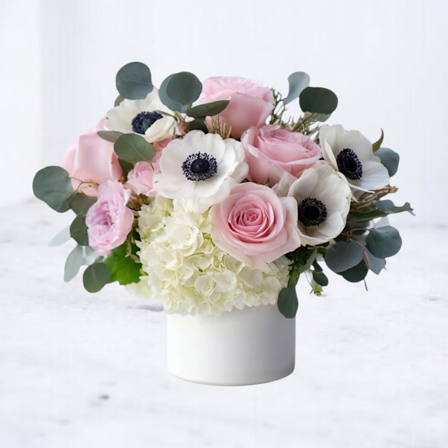 Anemone Alchemy - A compact, rounded bouquet of pink and white blooms that feels like a soft exhale - perfect as a birthday arrangement or a gentle "I'm sorry" gesture. The low, calming profile and romantic pastel palette make this flower arrangement a thoughtful Spring surprise or a quiet "thinking of you" gift that will help the person you care about feel seen and soothed.  Creamy white hydrangea forms a cushion-like base for soft pink roses and white anemones with dark centers provide striking contrast. The flowers are gathered tightly in a white cylinder vase for an understated, modern presentation that sits nicely on a dining table, bedside table, or office desk.  Whiskey Wallflower is a design-forward florist serving Magnolia, The Woodlands, Montgomery, Willis, Conroe, Spring, and surrounding Houston-area communities. We specialize in thoughtfully composed, modern floral arrangements with a focus on premium blooms, intentional design, and reliable same-day delivery. Every piece is created with care to make life’s moments feel elevated, personal, and unforgettable.