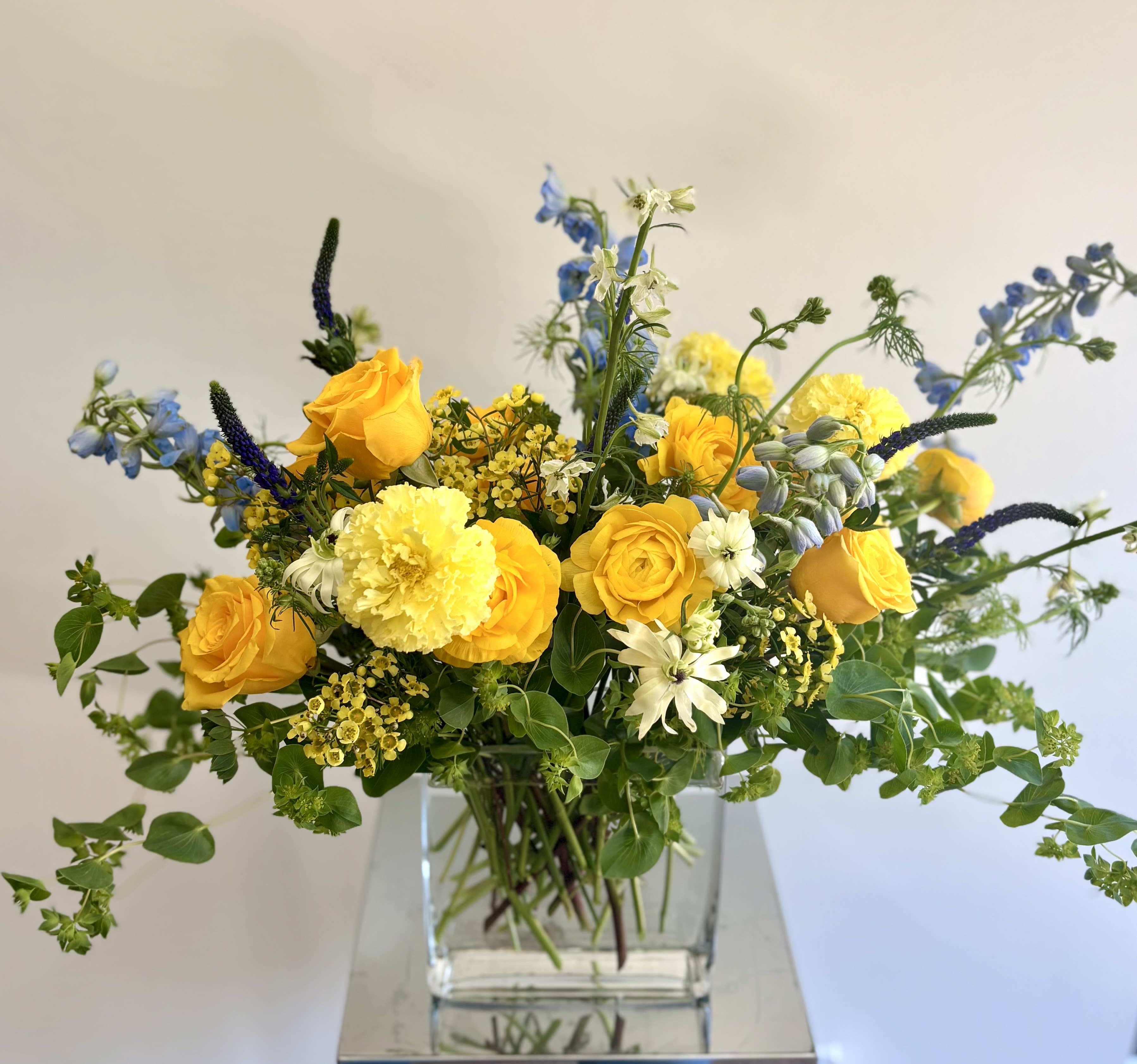 Melinda - Lovely garden-style flower arrangement with blue-yellow-white blooms in a glass vase. Michaela carefully selects the freshest flowers with the mix of roses, lisianthus, delphinum, ranunculus, tulips when available, bupleurum and other premium flowers and fillers to make this flower arrangement sunny and happy.