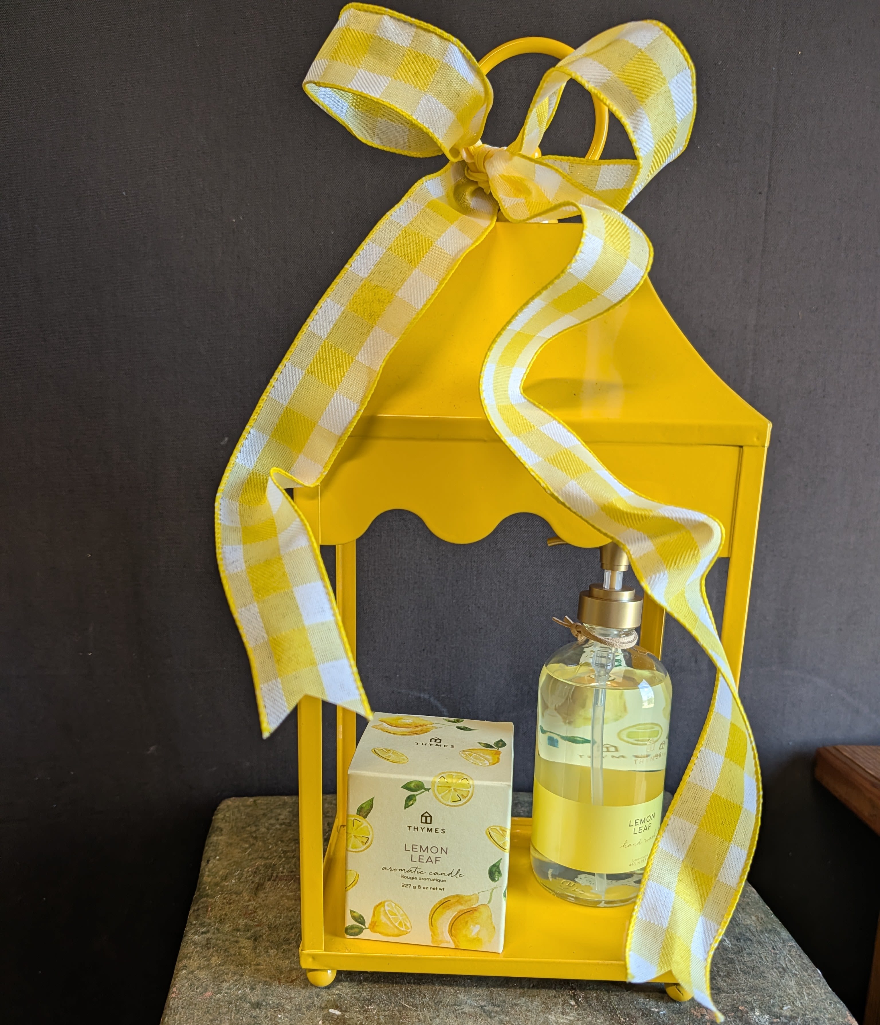 Lemon Twist Lantern Gift Set - Sunny citrus cheer arrives in a Lemon Twist Lantern gift set. An uplifting gift set perfect for birthdays, housewarmings, thank-you gestures, or a bright pick-me-up. The vivid yellow metal lantern and large yellow-and-white plaid bow make an immediate visual impact, so the person you're thinking of will feel noticed and refreshed the moment they see it.  Inside the lantern a boxed Thymes lemon leaf candle with illustrated lemons sits beside a Thymes lemon leaf clear glass hand wash pump bottle, creating a coordinated lemon-themed display in yellow accents. The ribbon's long trailing ends frame the contents and add a tactile, giftlike finish; the compact lantern is ready to display on an entry table, kitchen counter, or mantel for instant cheerful décor.  This set is designed to be both decorative and fragrant-ideal for welcoming someone into a new home or saying thanks in a way that brightens their day. Order with standard delivery and the lantern arrives assembled and gift-ready so the recipient can enjoy the scent and the sunny styling straight away.