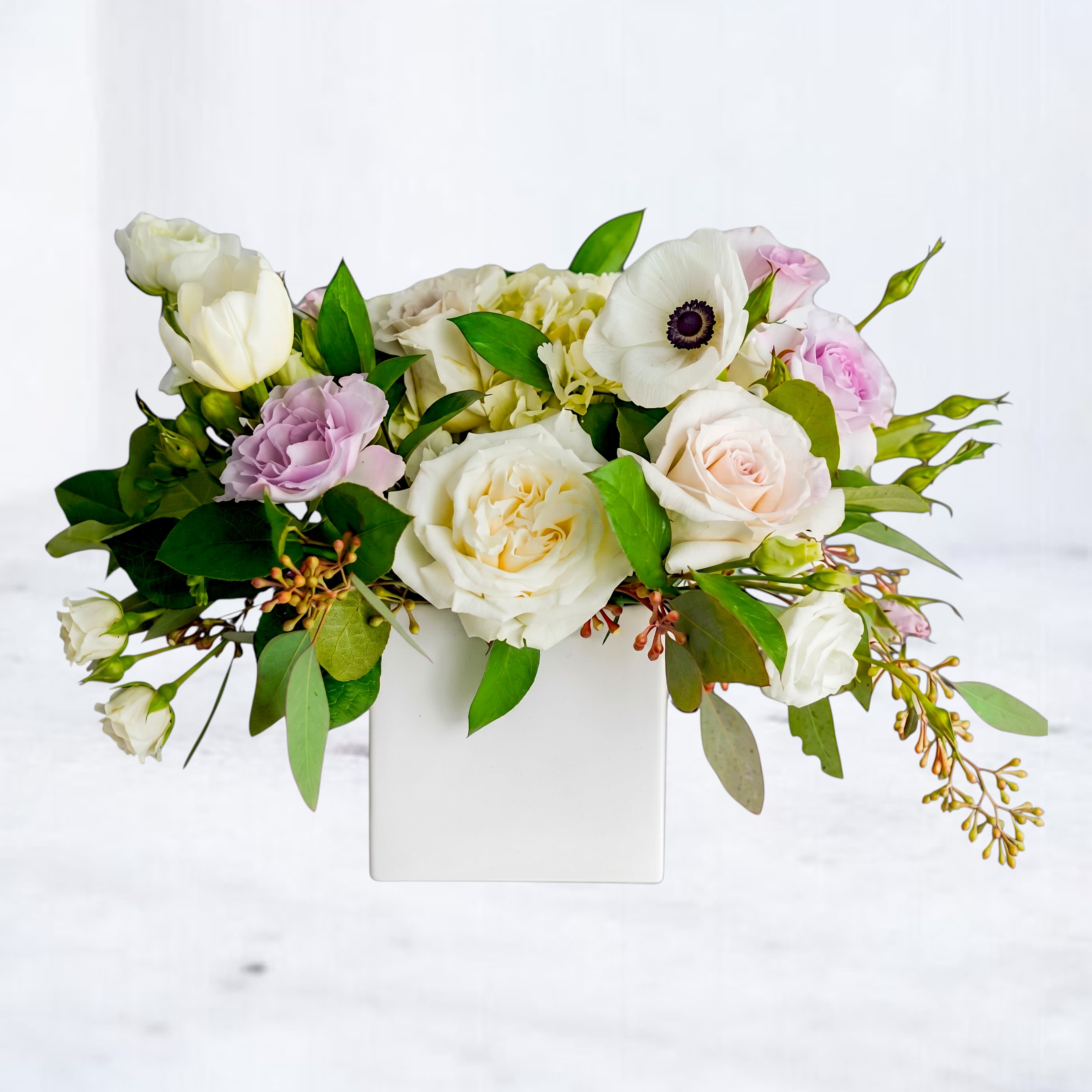 Botanist - Soft white and blush blooms arranged low in a white square vase create a gentle, garden-fresh impression perfect for a sympathy or thinking-of-you bouquet. This flower arrangement for "just because" moments pairs large white roses with pale pink roses and soft lavender lisianthus so the person you're thinking of will feel comforted and quietly celebrated rather than overwhelmed.  The composition includes a single white anemone with a dark center, a pale hydrangea cluster, spray roses and additional mixed blooms tucked among abundant greenery and trailing accent stems for an airy, romantic silhouette. Presented in a clean white square vase, the design sits wide and low-easy to display on a mantel or bedside table-and reads as refined and natural across seasons.  Ideal when you want to say you care without words, whether for a funeral, to offer condolences, or simply to brighten someone's day. Arrives ready-to-display in the included vase; keep in fresh water, trim stems every few days, and enjoy for as long as the blooms last.