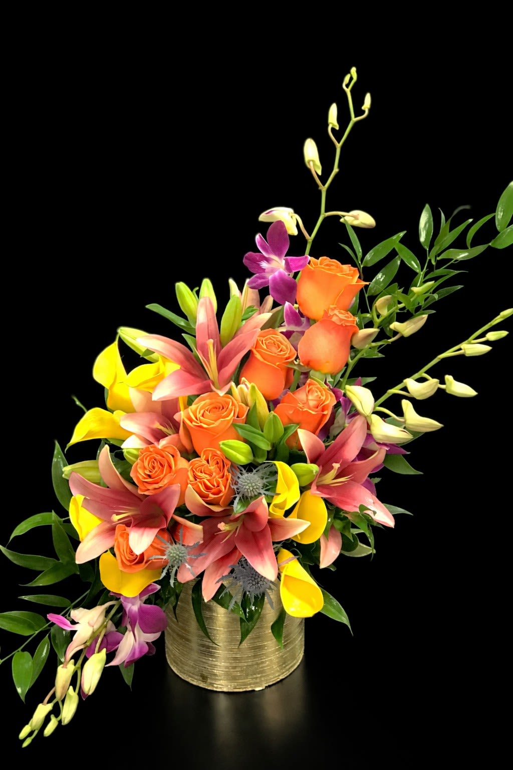 Orange roses with pink lilies and yellow calla lilies in a gold vase