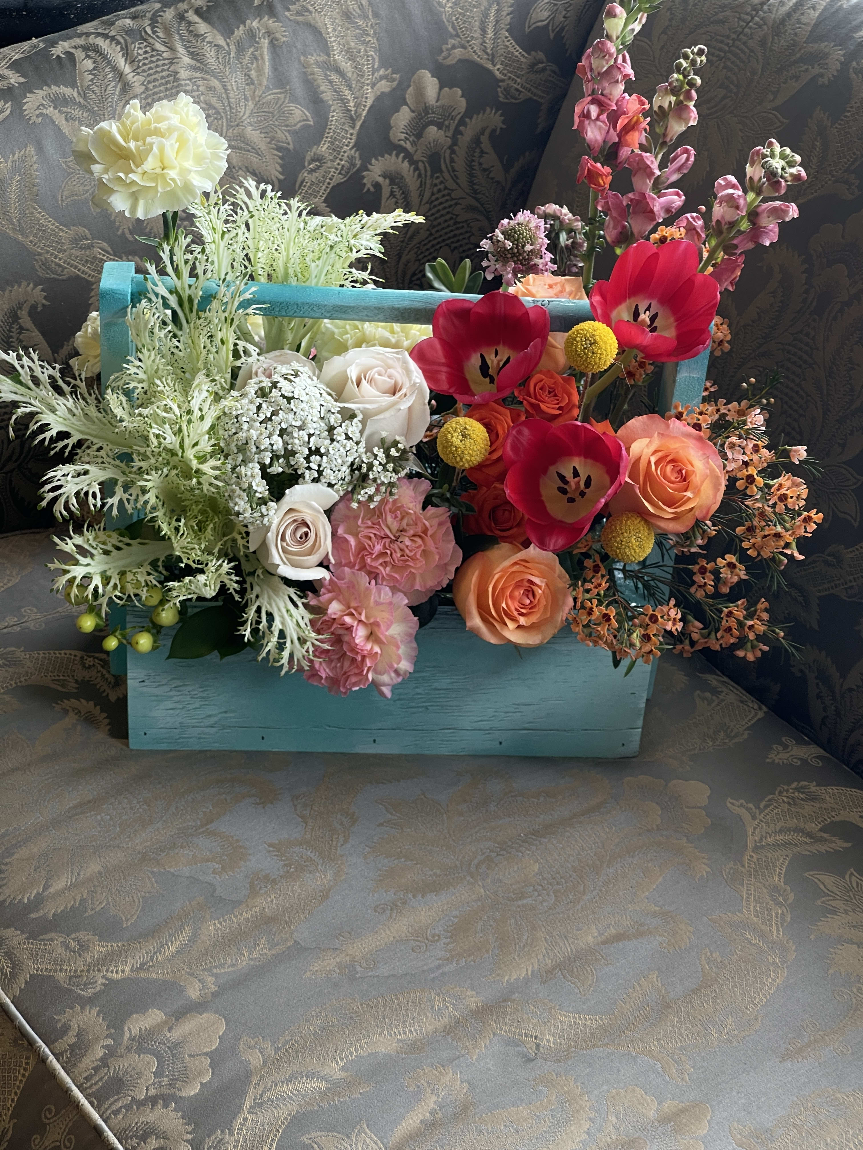 Designers Choice in English Garden Style  - A sunlit mix of garden roses, gerbera-style blooms and playful accents arrives in a turquoise wooden box with a handy handle - a lively flower arrangement for Mother's Day, birthdays, anniversaries or simply "just because." The English garden-inspired design feels casually elegant: soft blush and cream roses and pink carnations sit alongside hot pink tulips and orange roses, while tall pink snapdragons and yellow billy balls add height and cheerful contrast that will make the recipient feel warmly celebrated.  This mixed bouquet includes garden roses, roses, carnations, tulips, snapdragons, stock, gerbera-style flowers, small orange clusters, delicate white filler blooms and a small succulent tucked into the center. The asymmetrical, spilling composition gives a fresh, hand-gathered look, and the turquoise wooden box with handle lends rustic charm and tabletop-ready display - no vase required. Ideal for occasions from birthdays, graduations, and get-well wishes to Thanksgiving, Christmas, sympathy or a thoughtful thank-you, it brings vibrant color and texture to every season; size and exact flowers may vary slightly with seasonal availability.