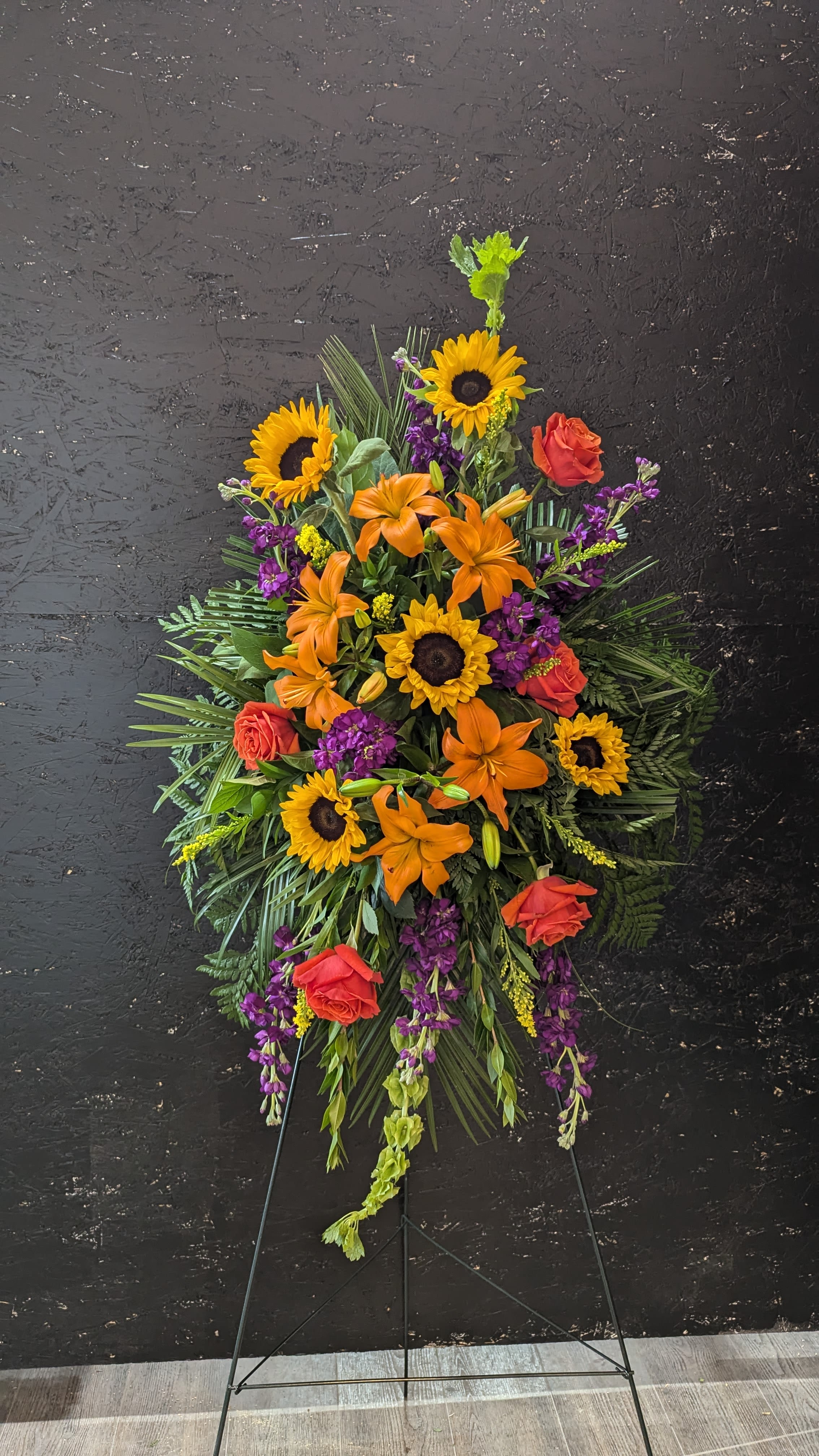 Sunset Serenity - Standing spray includes, Orange Lilies, Sunflowers, Orange Roses, Violet Stock, Bells of Ireland and assorted greenery.