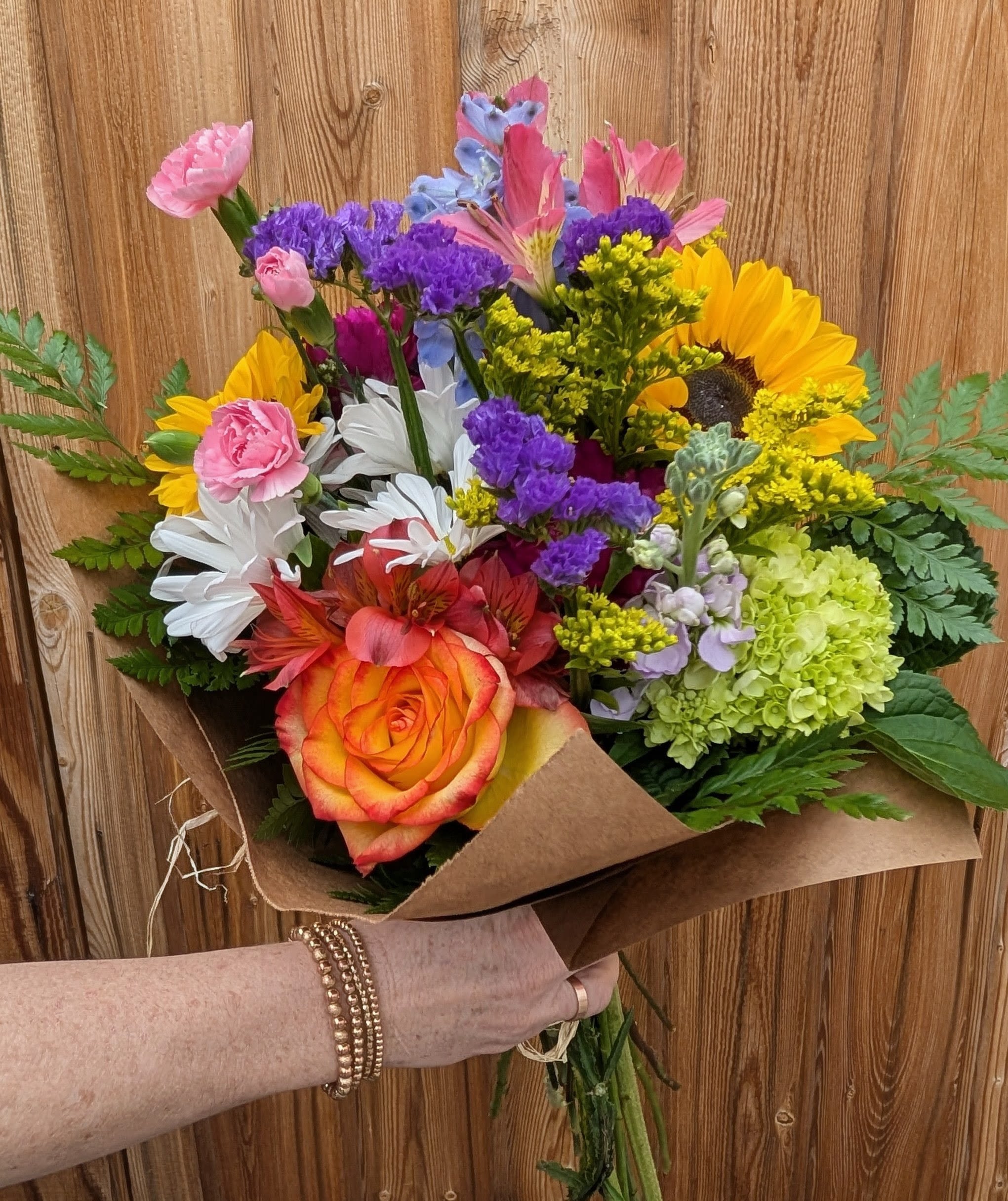 Small Hand Tie - A bright, market-style hand-tied bouquet that feels like sunshine in brown paper-the Small Hand Tie is a cheerful mixed flower arrangement perfect for birthdays, thank-you gifts, or just because. The first impression is the large orange-and-yellow rose paired with bold yellow sunflowers, so the person you're thinking of will feel noticed and celebrated the moment they see it. 