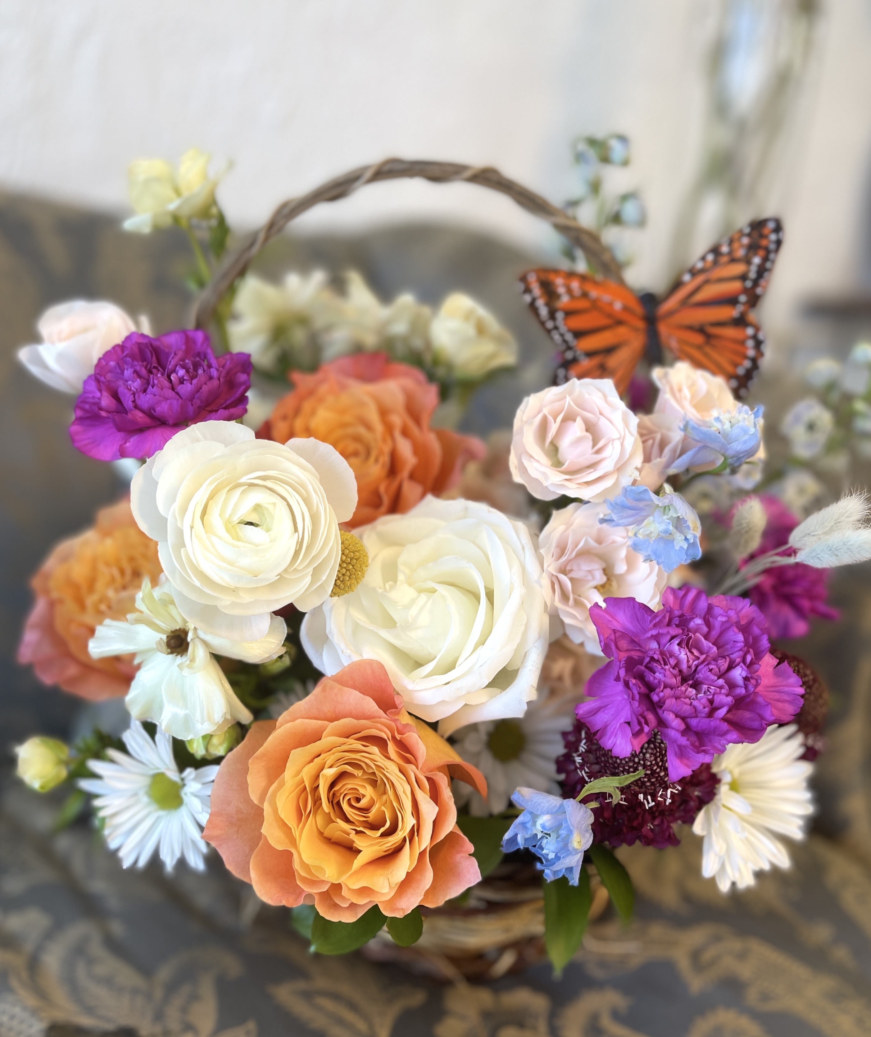 Basket arrangement of roses, daisies, and purple blooms with a butterfly accent