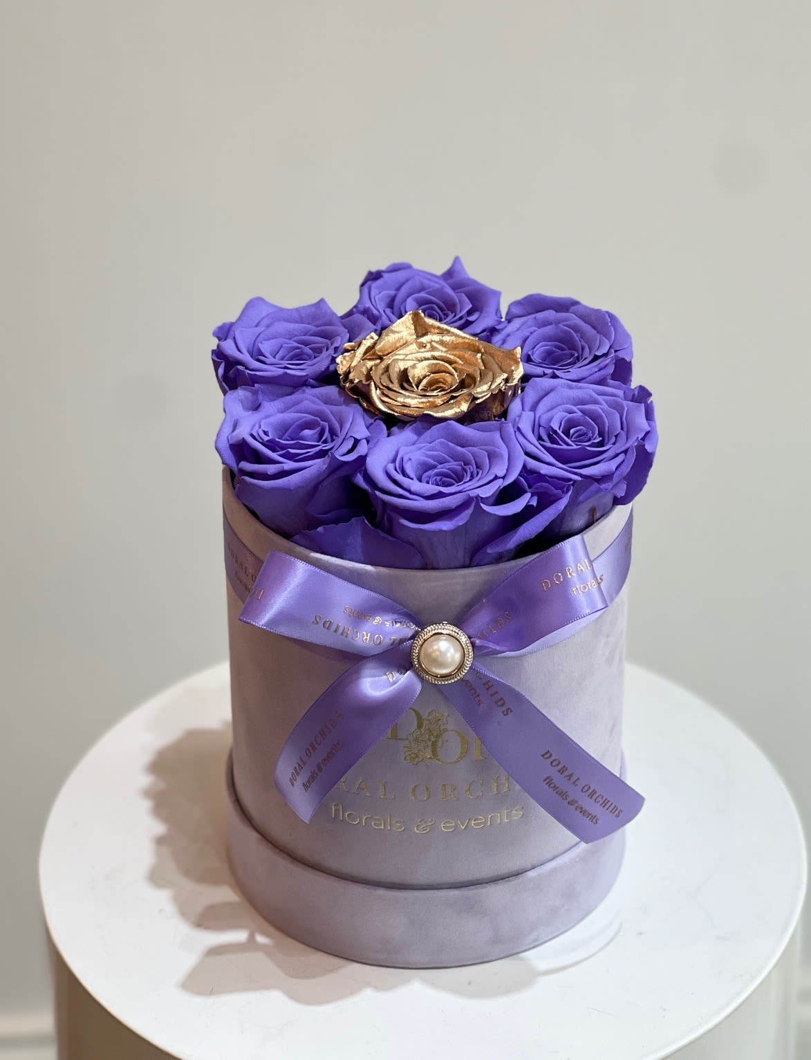 Purple roses in a round lavender box with a gold rose center and ribbon bow