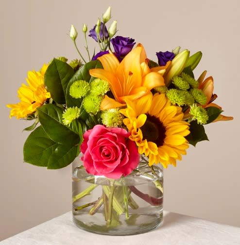 Mixed bouquet with sunflowers, roses, lilies, and purple blooms in a glass vase