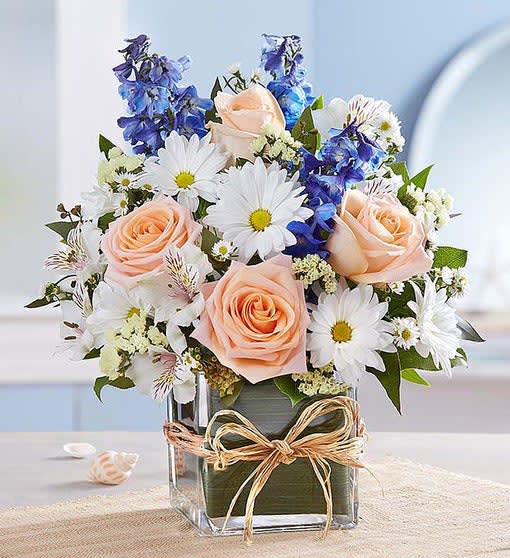 Peach roses and white daisies in a clear square vase with a ribbon