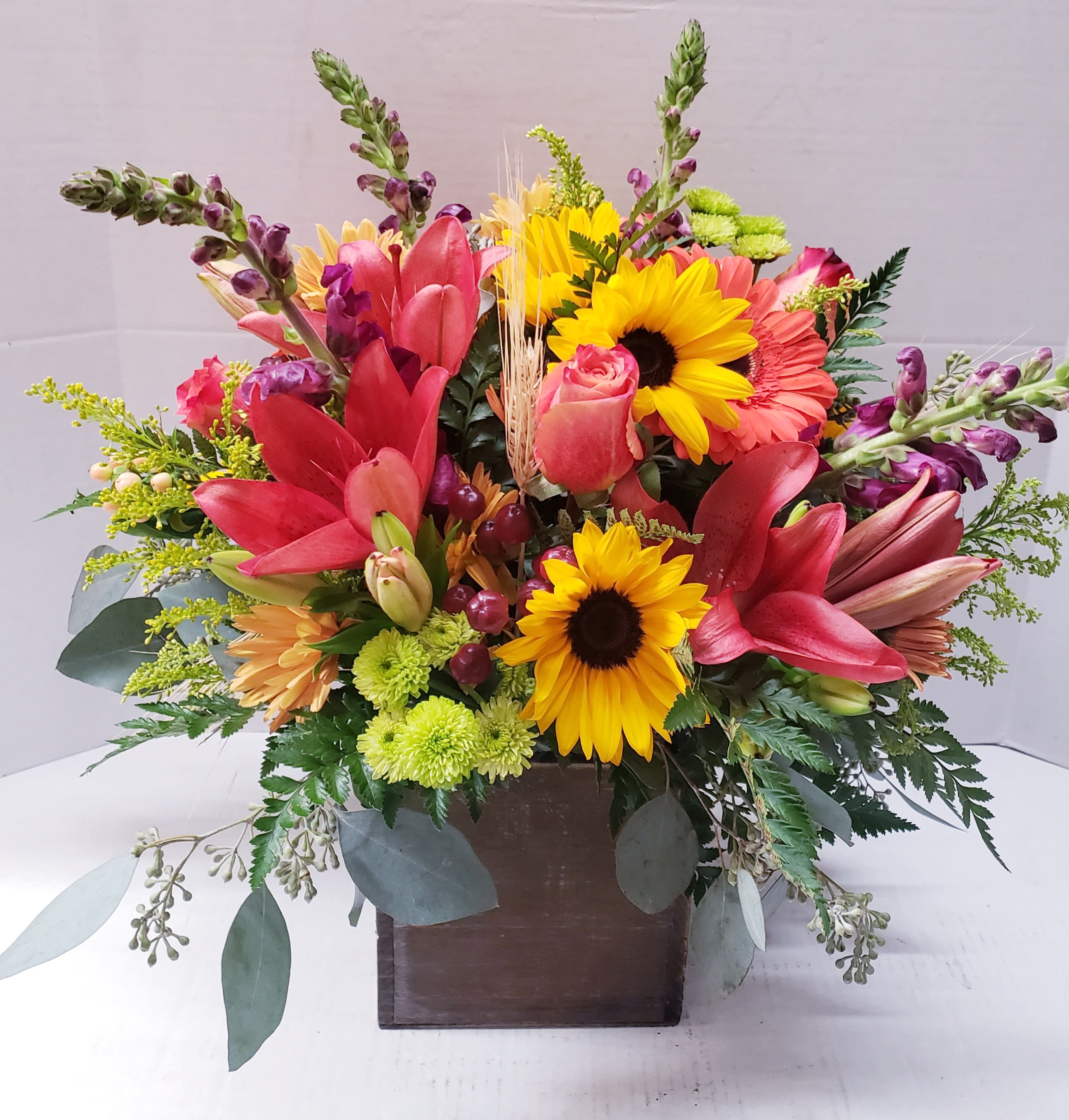 Bright mixed arrangement with red lilies, yellow sunflowers, and roses in a square wooden vase