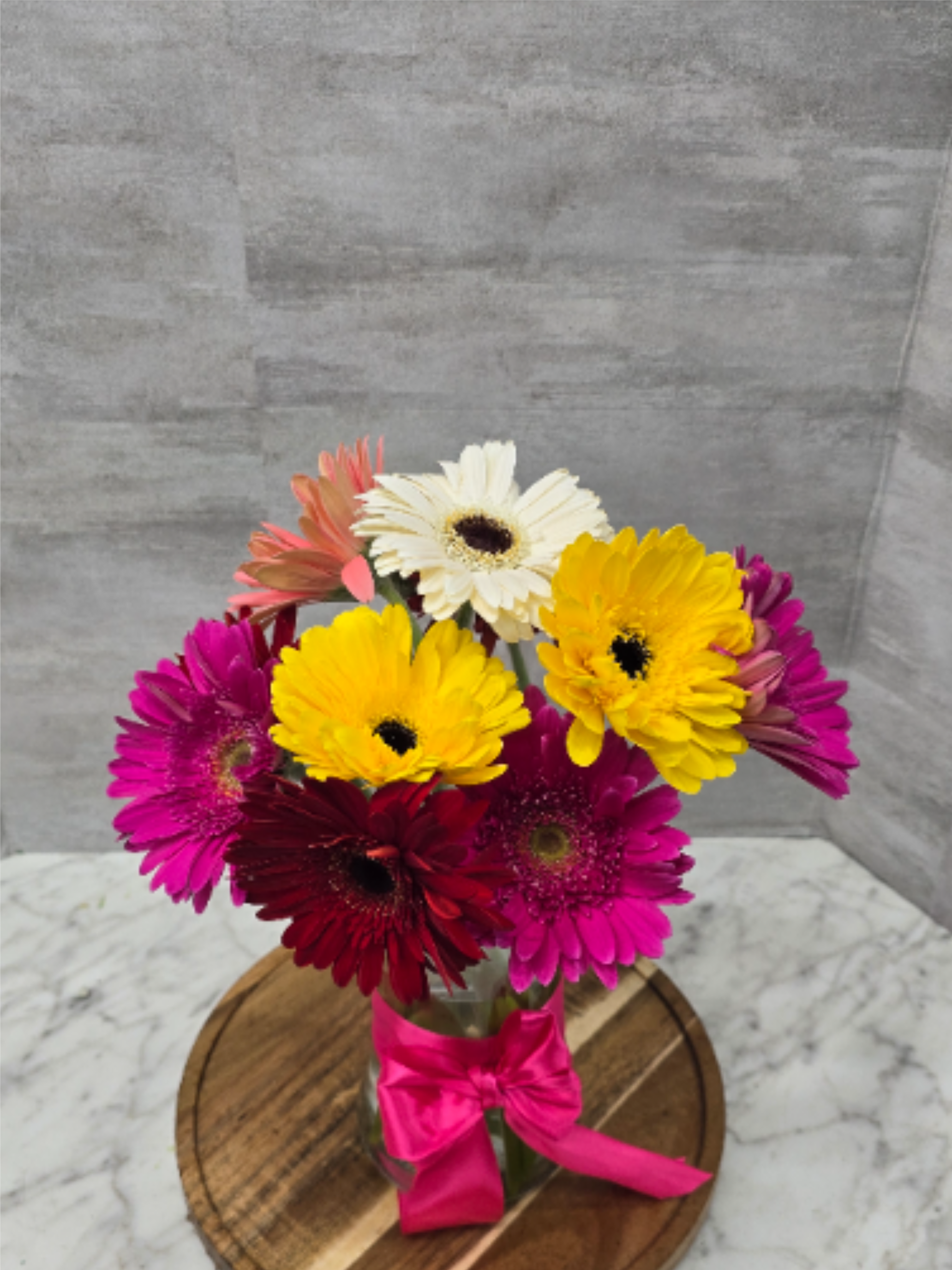 Gerbera Surprise - A burst of cheerful color-Gerberas come together in a compact, playful cluster that's perfect for a birthday bouquet, congratulations gift, or a spontaneous "just because" surprise. The arrangement reads lively and upbeat; the person you're celebrating will feel genuinely noticed and uplifted by the saturated tones and friendly, open faces of the gerberas.  Tightly gathered stems sit in a clear glass vase wrapped with a vivid pink satin ribbon and are presented on a simple wooden round tray for easy display. The compact size makes it right for tabletops, desks, or a kitchen counter; the sturdy glass and tray give it an instant, polished look without fuss. No special care is needed beyond fresh water and a cool spot away from direct sun to keep the blooms looking their happiest for days.