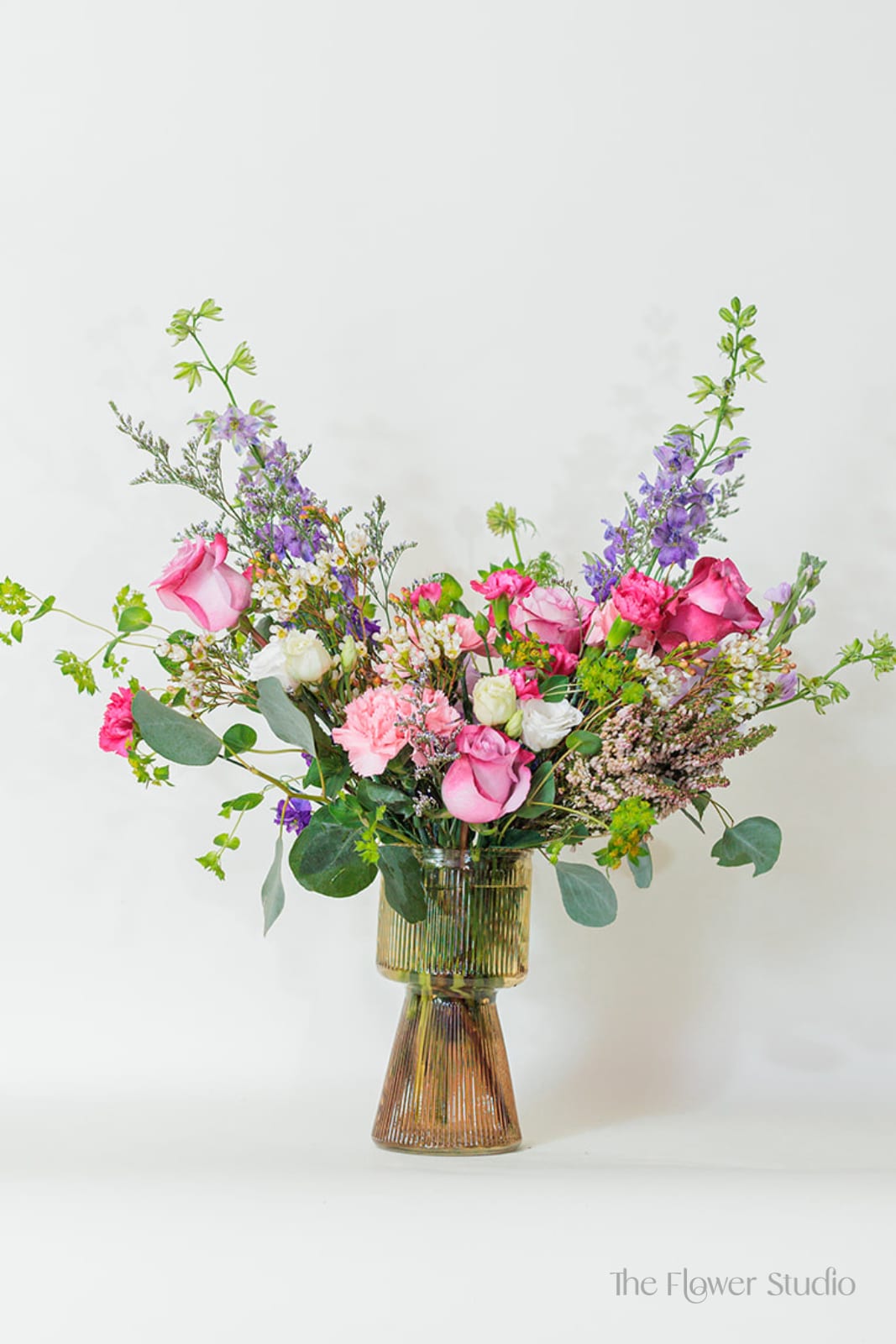 Tall mixed arrangement of pink roses, carnations, and purple flowers in a ribbed glass vase