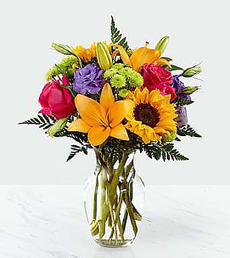Colorful bouquet with sunflowers, lilies, roses, and purple blooms in a clear glass vase