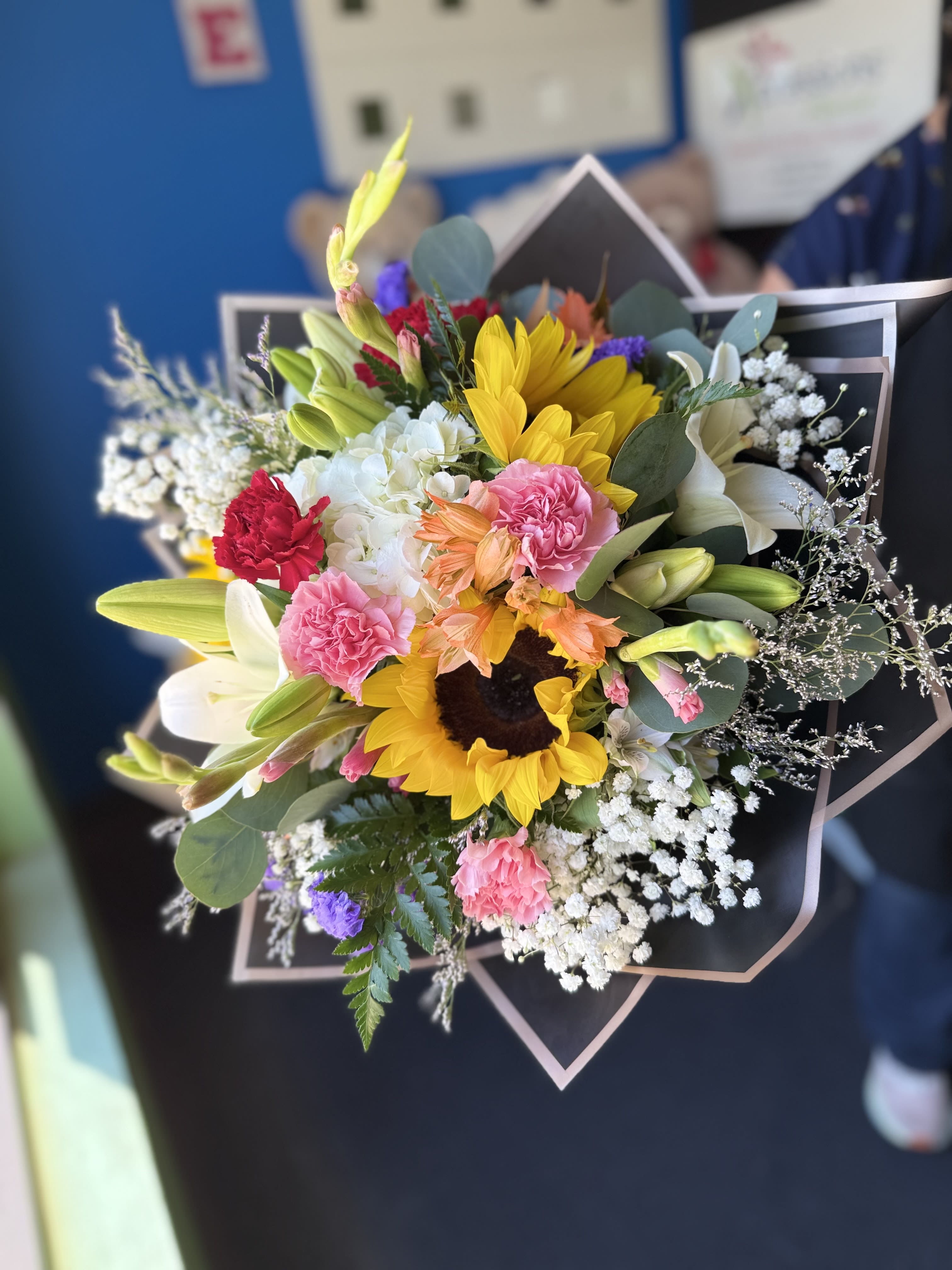 Spring Fever Bouquet - (WRAP-NO VASE) Spring Fever Bouquet is a vibrant and cheerful arrangement bursting with seasonal blooms like sunflowers, lilies, carnations, and delicate fillers. Designed to capture the beauty of spring, this bouquet features a fresh mix of colors and textures. Each arrangement is unique