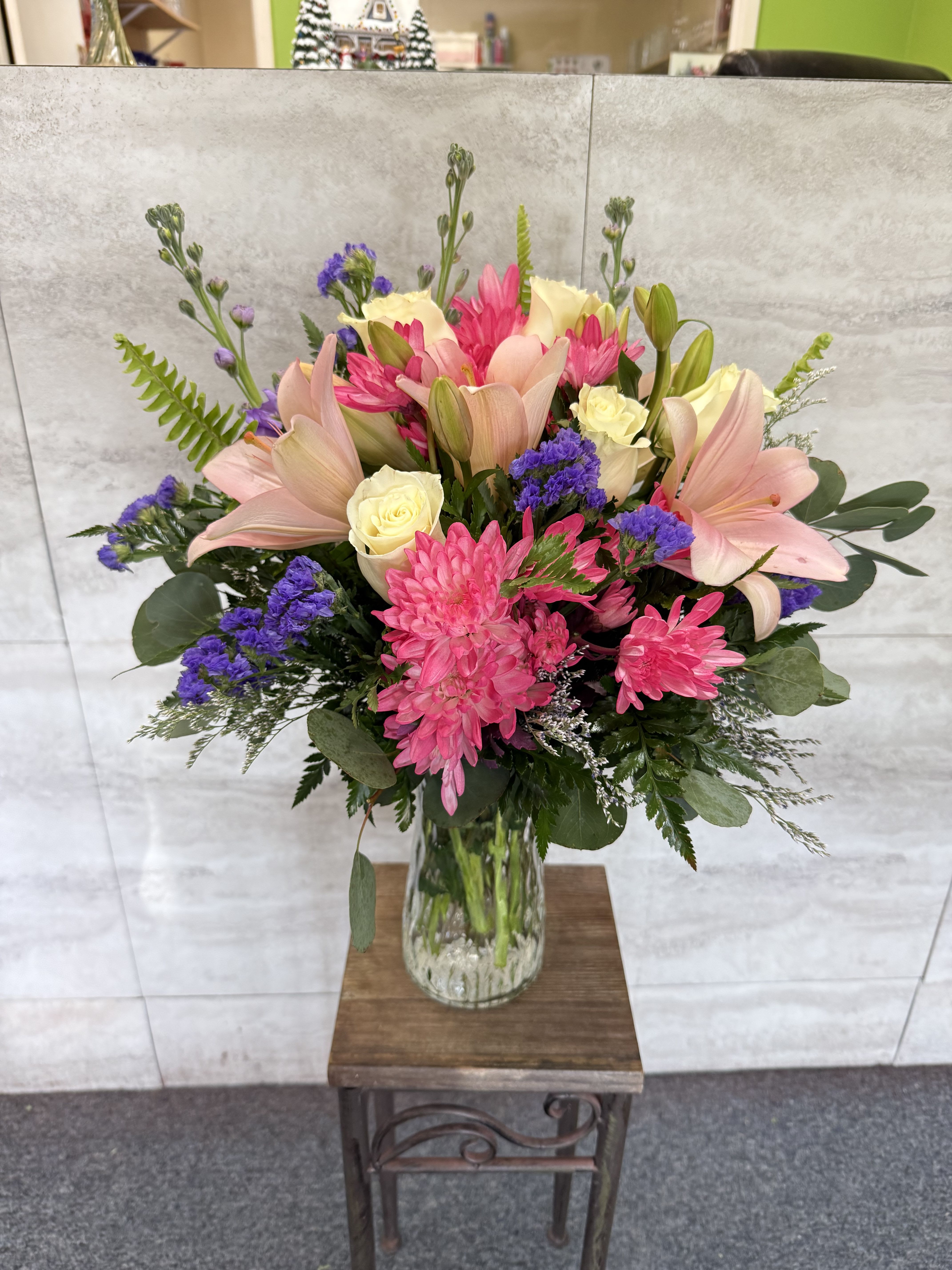 Soft Kisses - (ALL-ROUND) This vibrant and lush floral arrangement features a mix of soft pastels and bold pops of color. The bouquet showcases elegant light pink lilies and crisp white roses, beautifully contrasted by bright pink chrysanthemums and dainty purple statice.  Deep green ferns and eucalyptus leaves add texture and fullness, all presented in a classic clear glass vase. It’s a cheerful and balanced design that works well for birthdays, thank-yous, or everyday decor.
