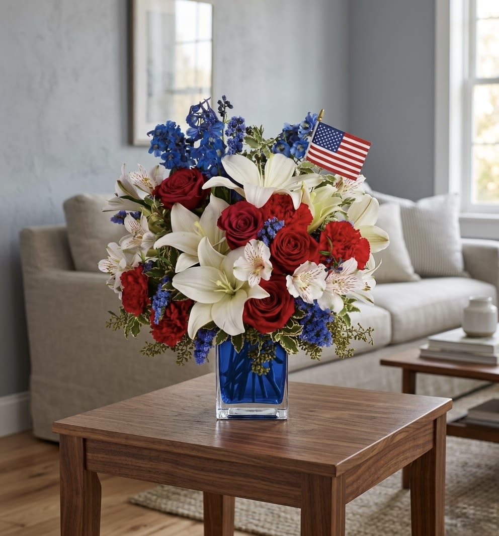 America The Beautiful  - Red roses, white lilies, red carnations and pale alstroemeria with vivid blue accents in a blue glass vase, topped with a small U.S. flag - bright, festive, and patriotic.