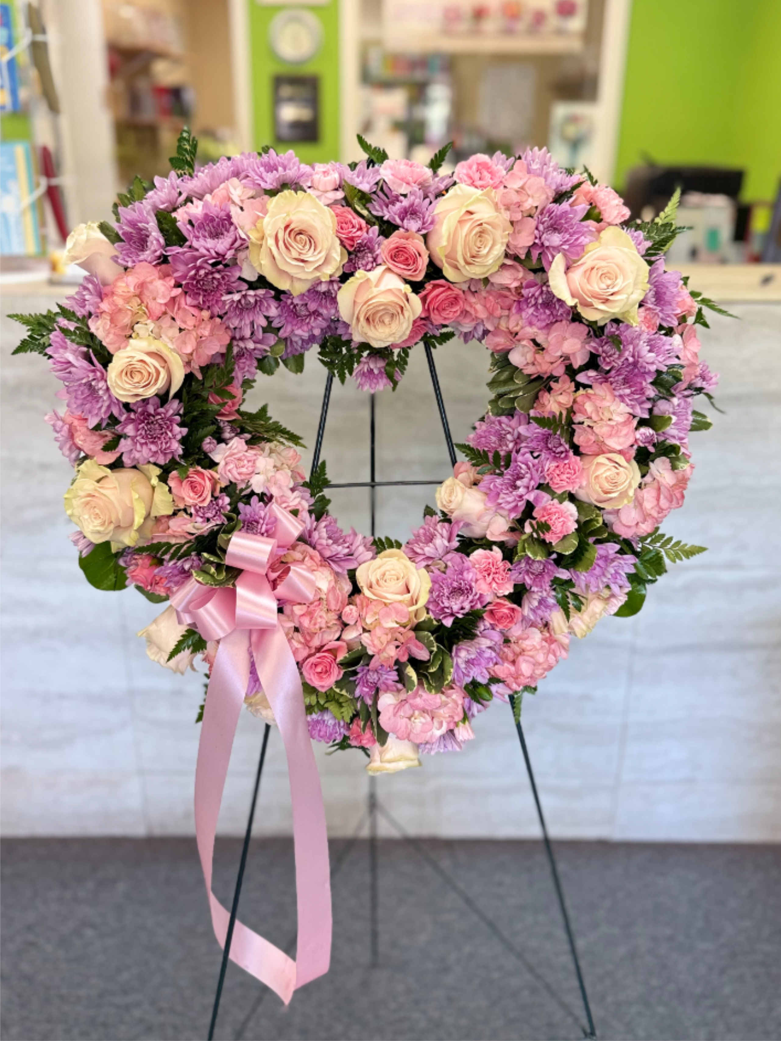Heart-shaped standing wreath of pink and lavender flowers with a pink ribbon bow on a metal easel