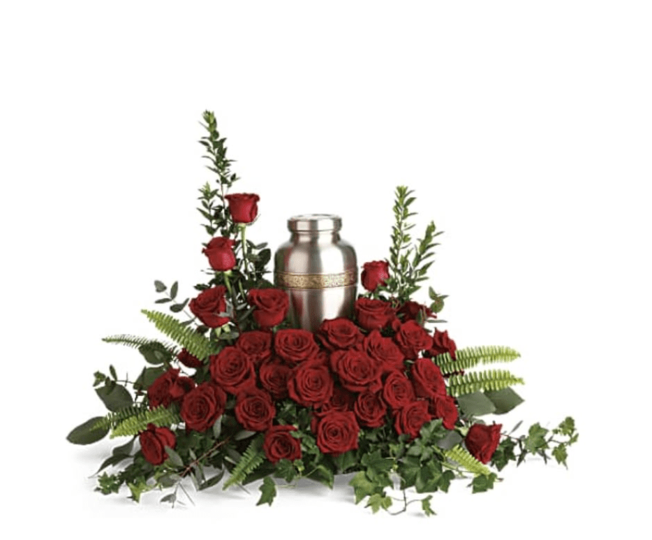 Forever In Our Hearts Cremation Tribute - A bed of deep red roses cradles a silver-toned metal cremation urn, creating a solemn and dignified sympathy arrangement that honors a life with quiet elegance. This funeral flower arrangement features a low, elongated shape with a few slightly taller rose stems at the back to gently frame the urn, making it an appropriate choice for memorial services, cremation ceremonies, or any moment of lasting remembrance.  The arrangement pairs rich, velvety red roses with lush green foliage and ferns that spill around the base, adding texture and contrast while drawing the eye to the central urn. Designed to sit on a casket stand or memorial table, the composition reads formal and reverent; the urn is shown for display context but is not included with the flowers. For families seeking a respectful expression of enduring love, this piece provides a concentrated, tasteful focal point for funeral and sympathy settings.