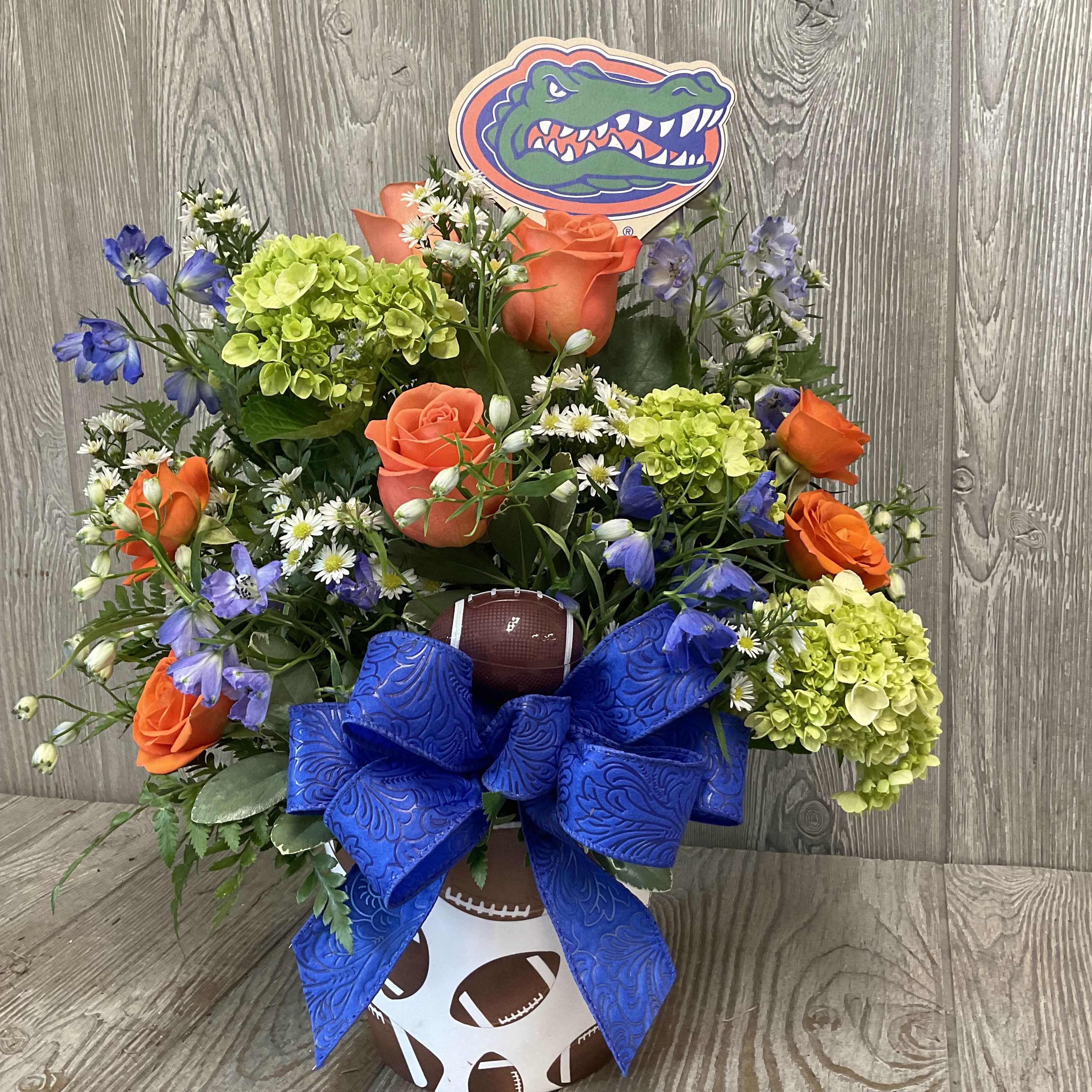 Football-themed bouquet with orange roses, green hydrangeas, and a blue bow in a sports container.