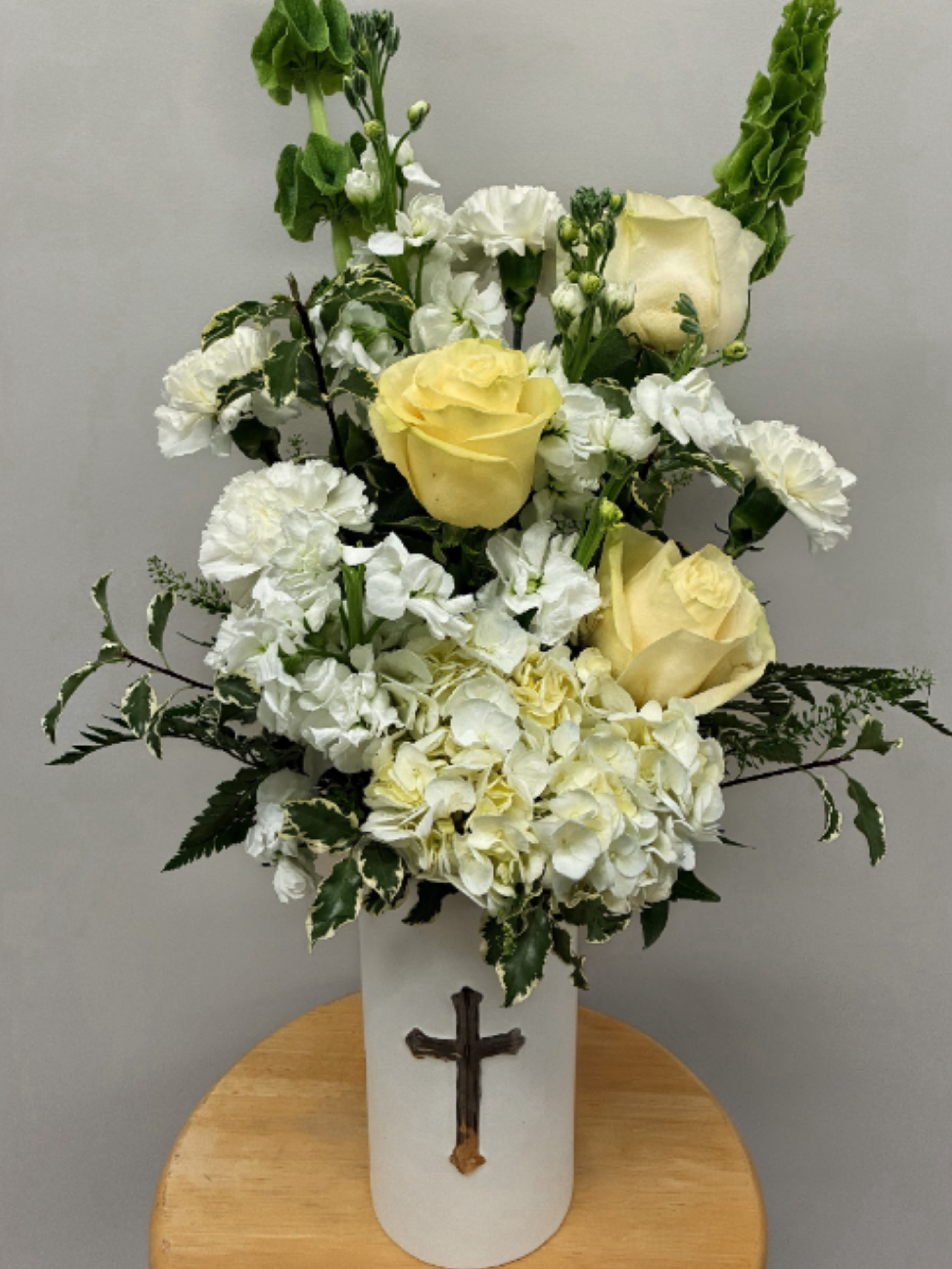Loving Tribute - Cream roses tucked among clusters of white carnations and a generous spray of white hydrangea give this sympathy arrangement a quietly uplifting presence. Designed as a memorial or church arrangement, the vertical green spikes and variegated foliage create an elegant, upright silhouette that reads reverent and composed-the kind of floral tribute that helps the person you're honoring feel remembered with grace.  Arranged in a tall white ceramic vase accented by a glossy silver cross, the piece combines rounded blossoms and linear greenery for contrast in texture and form. The soft white, cream palette keeps the focus gentle and respectful; carnations and hydrangea lend fullness while roses provide a warm, personal note.   Perfect for funerals, church services, or quiet home memorials, this arrangement offers a calm, devotional expression of sympathy and faith. The tall vase makes a stately statement on an altar or foyer table; simple care-fresh water and a cool spot away from direct sun-will help the flowers look their best through the service.
