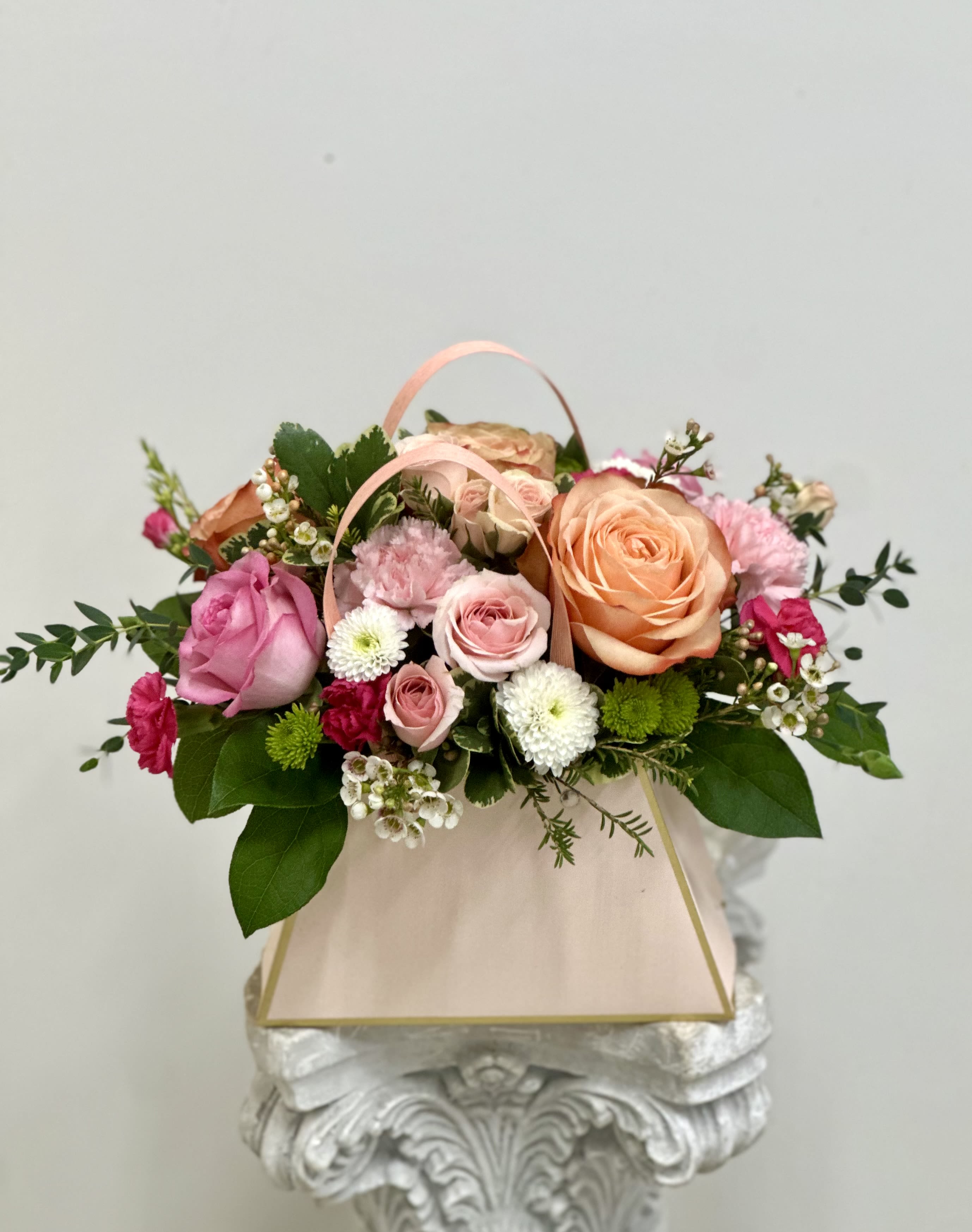 Pastel pink and peach flower arrangement with roses and mums in a blush gift bag-style container.