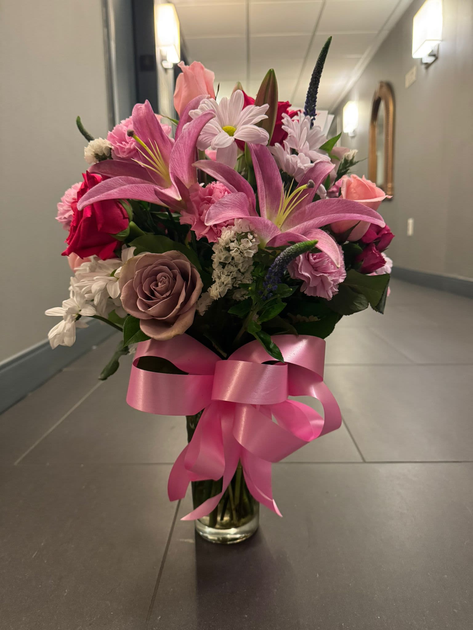 "Rosa Encantada" - Transform any moment into a fairy tale with Rosa Encantada.  This delicate and romantic arrangement combines the strength of pink lilies, the tenderness of mauve roses and the freshness of white daisies, all in a perfect balance of color and texture. The vibrant pink bow that embraces the vase symbolizes the union between classic elegance and modern style, making it the ideal detail to steal smiles.