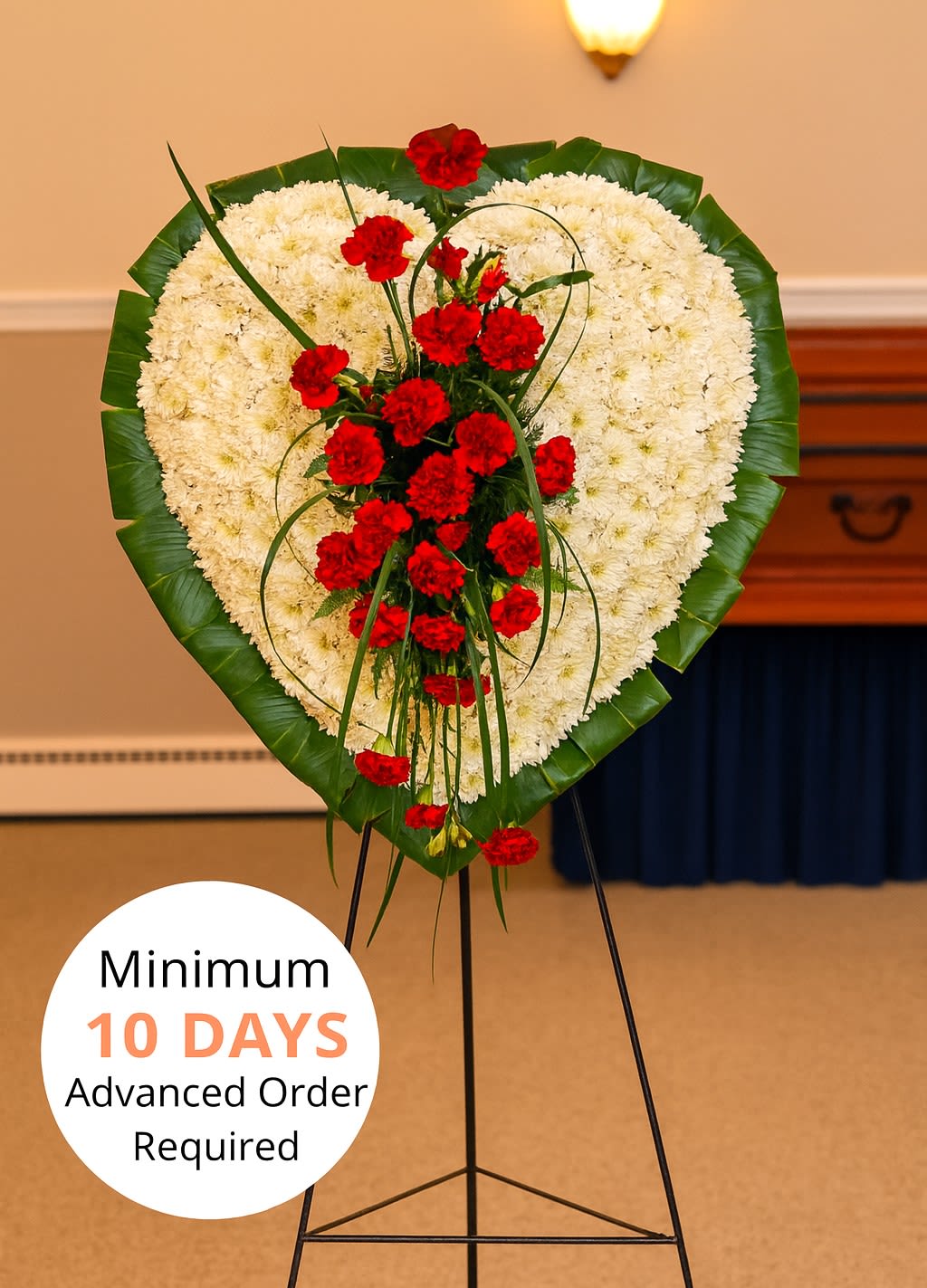 Heart-shaped standing wreath of white flowers with red carnations and a green leaf border on an easel.