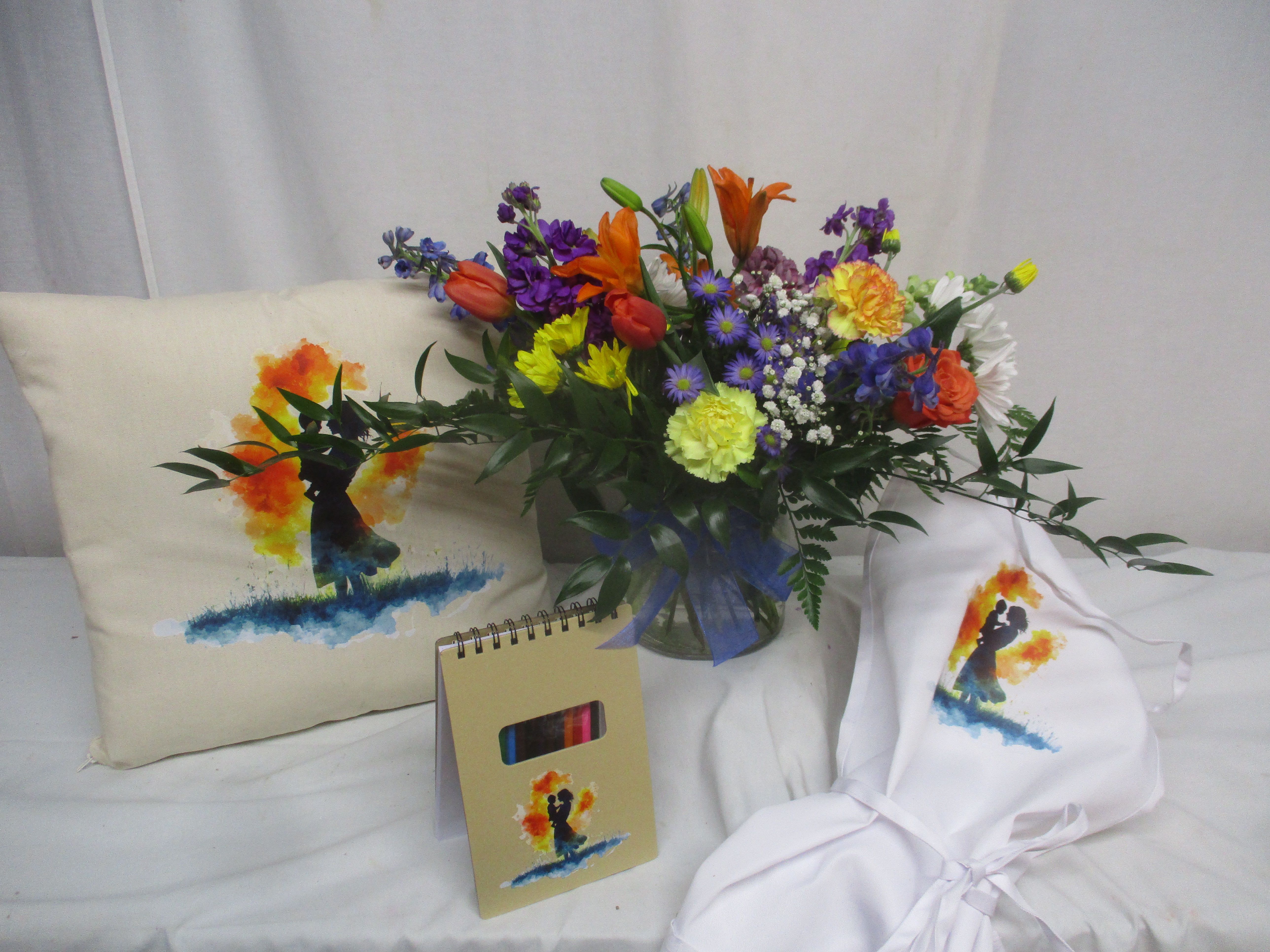 Beauty Bundle - Lg - Bright, multicolored tulips, carnations, daisies and lilies tumble from a clear glass vase in this Mother's Day bundle, creating a joyful spring bouquet perfect for Mother's Day, a birthday, or a "thinking of you" surprise. The lively mix of purple, orange, white, blue and warm yellow blooms-with soft greenery peeking through-gives the person you're celebrating an immediate lift; she'll feel seen and cherished the moment the arrangement arrives.  This large arrangement arrives as a coordinated gift set: the dense, loosely gathered bouquet is tied with a blue ribbon and presented alongside a cream decorative throw pillow printed with a tender mother-and-child silhouette, a matching spiral notebook and a white imprinted apron. The pillow's watercolor bursts in orange, yellow and blue echo the flowers' palette for a thoughtful, keepsake-ready presentation. The vase, pillow and stationery make this an all-in-one gift that suits home display, everyday celebration, or a cozy keepsake corner.  Designed for gifting ease, this bundle arrives ready to present-no extra wrapping required-so the recipient can enjoy the floral fragrance and keep the printed pillow and notebook as a lasting memento. The arrangement's size and full mix of seasonal flowers create a bold visual impact; place the vase out of direct sunlight and add water every day to help blooms last.