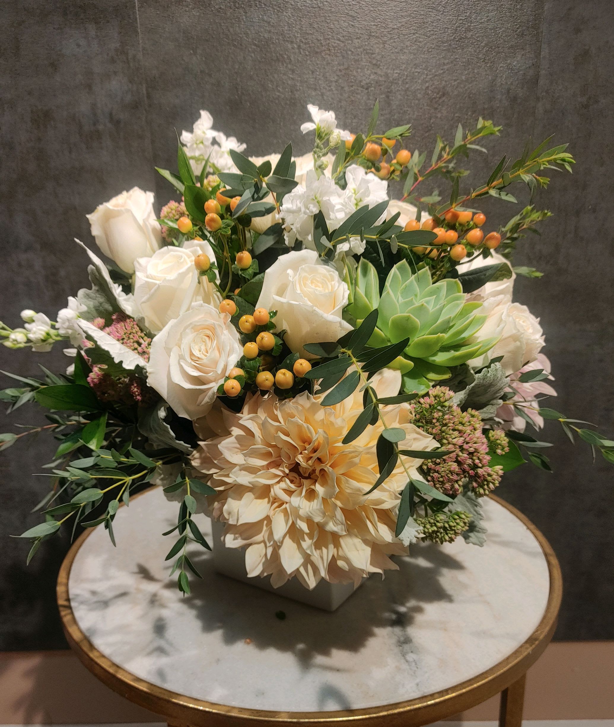 White roses and a peach dahlia arranged in a square vase with greenery.
