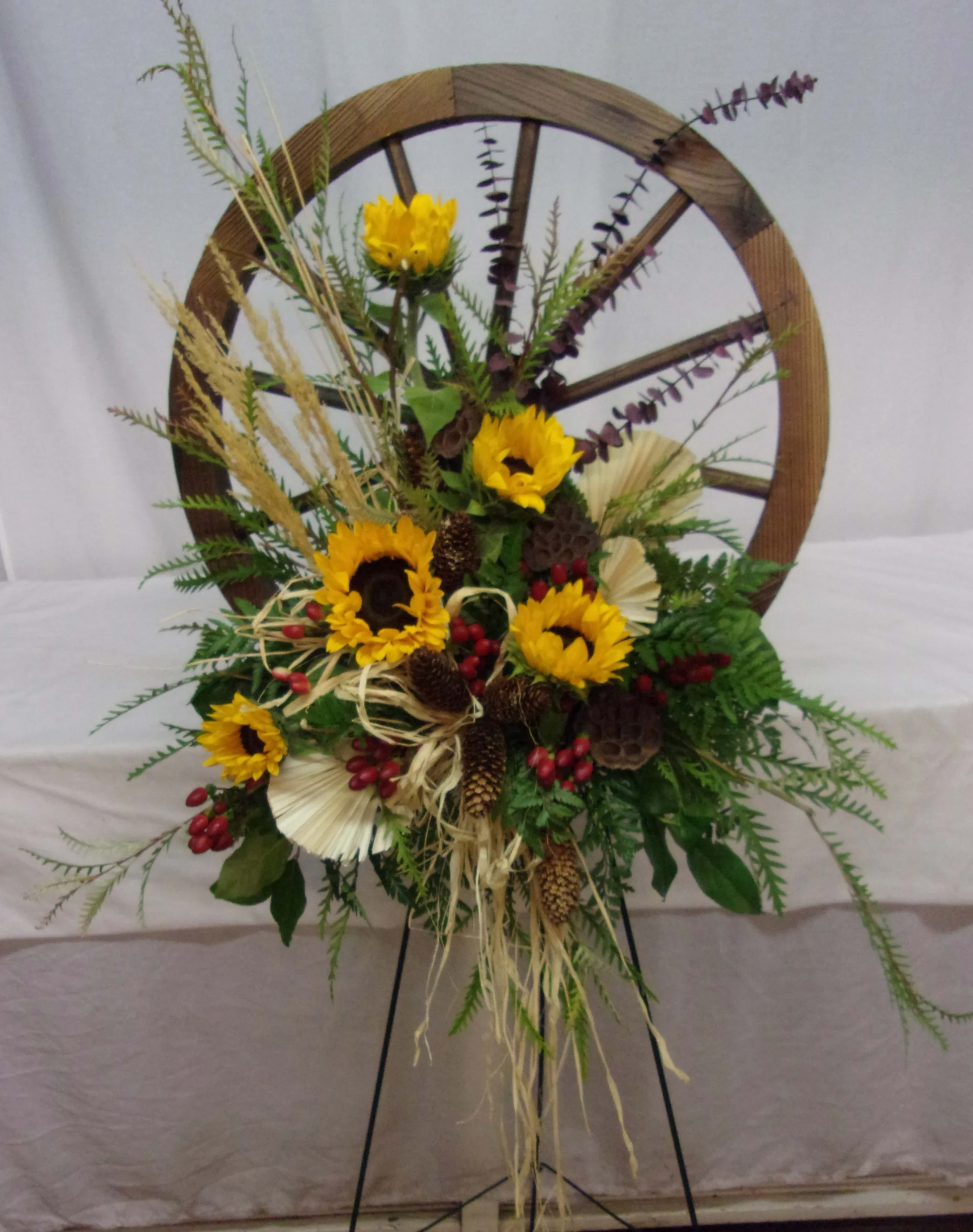 Sunshine Wagon Wheel  - A western style arrangement for the eternal cowboy, featuring a wooden wagon wheel with a floral display of stylish outdoor blooms.  **As many products contained in arrangement are natural, there will be variations from items as pictured.**