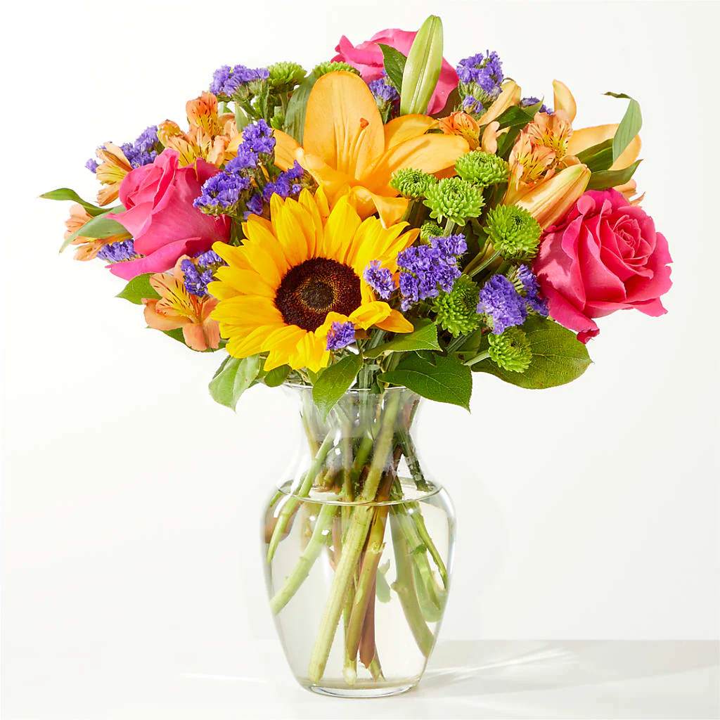 Mixed bouquet of sunflowers, roses, and lilies in a clear glass vase