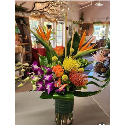 Aloha Dreams - Bright, sculptural and unapologetically exotic - this tropical flower arrangement is a bold birthday bouquet or "just because" surprise that reads like a celebration. Aloha Dreams brings orange birds of paradise, purple orchids and orange roses together with vivid yellow textured blooms so the person you're thinking of will feel seen, celebrated, and delighted by the vibrant color story and dramatic silhouette.  The design sits in a clear glass vase wrapped in a dark green decorative ribbon for a polished finish. Long arching leaves and broad tropical foliage form a wide, energetic shape that makes a striking centerpiece or gift for love and romance, congratulations, or any moment that calls for something memorable and luxe. Sized to command attention, it arrives ready-to-display with water in the vase; refresh water every few days and keep away from direct heat to maintain bloom life.
