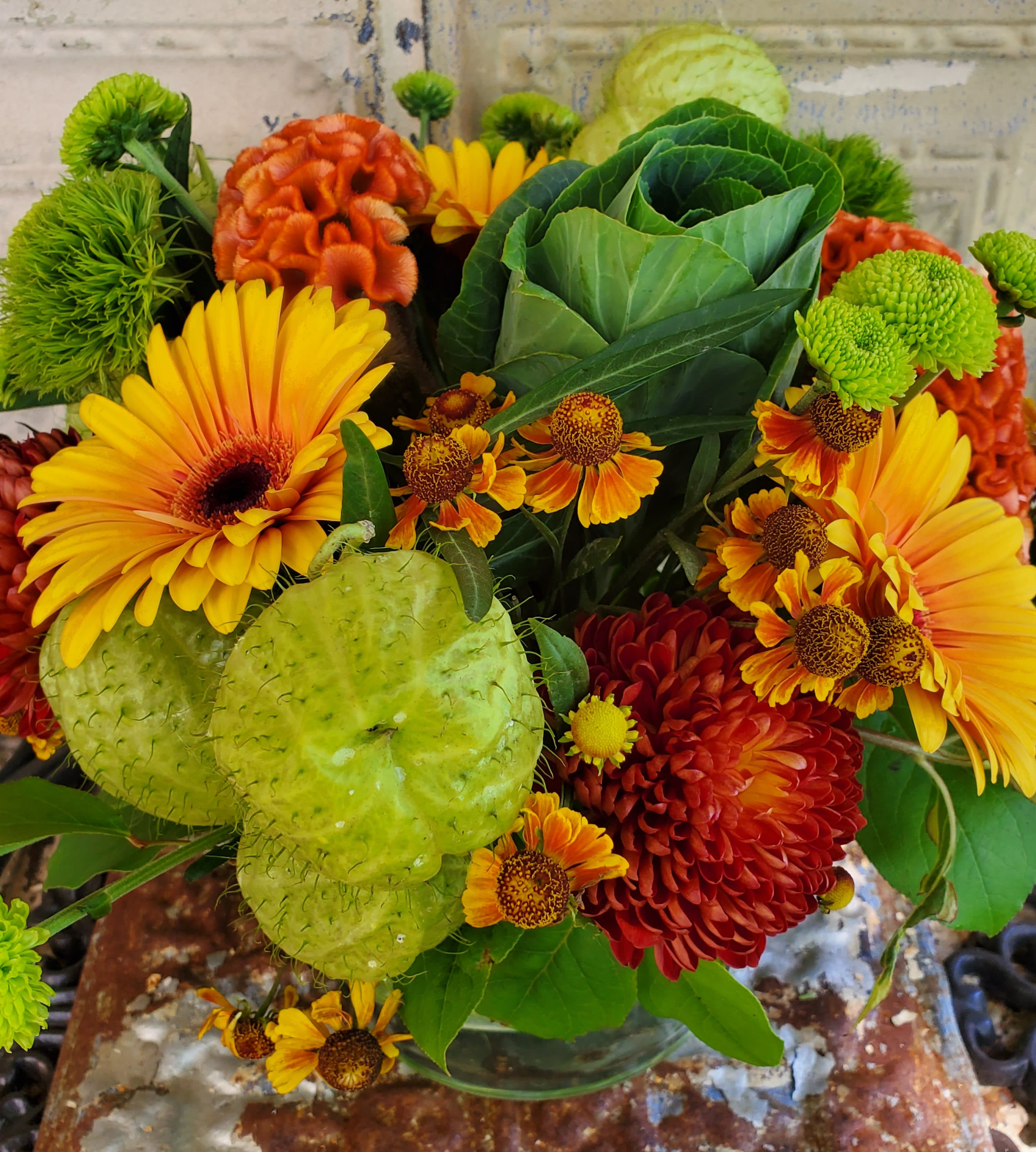 Bouquet of yellow gerbera daisies, orange mums, and green ornamental pods in a glass vase