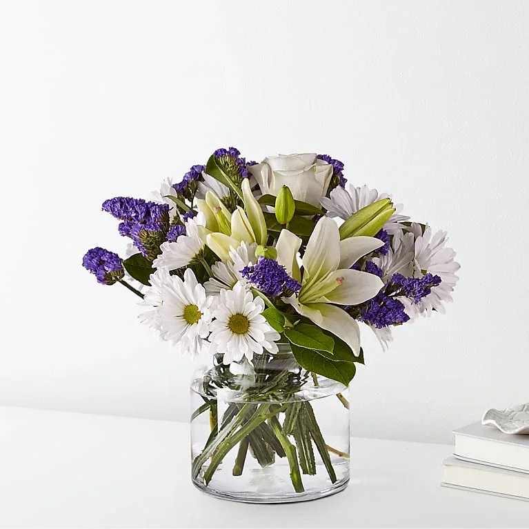 White lilies and daisies with purple filler in a clear glass vase