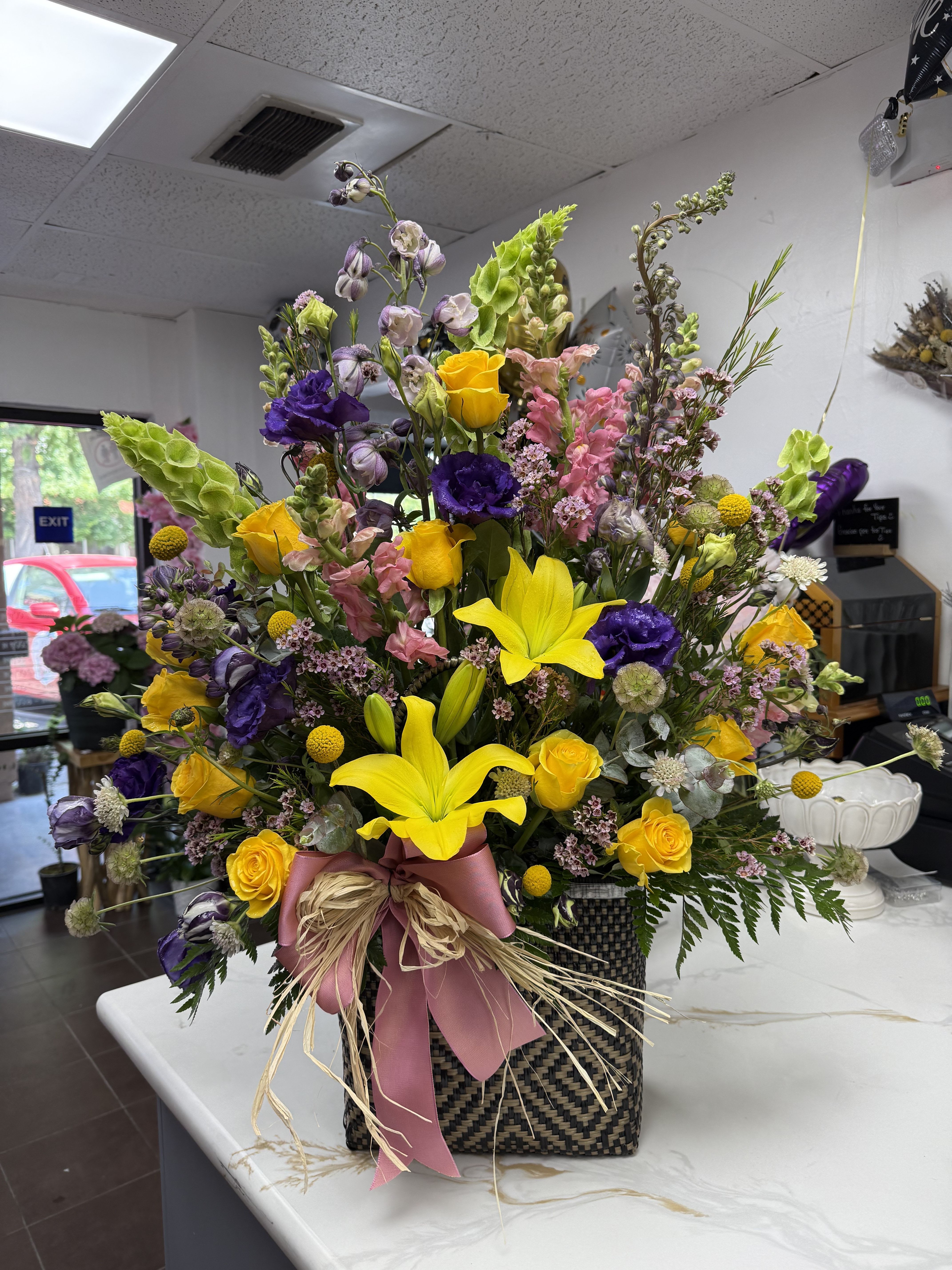 Tall mixed bouquet in a woven basket with pink ribbon and yellow lilies