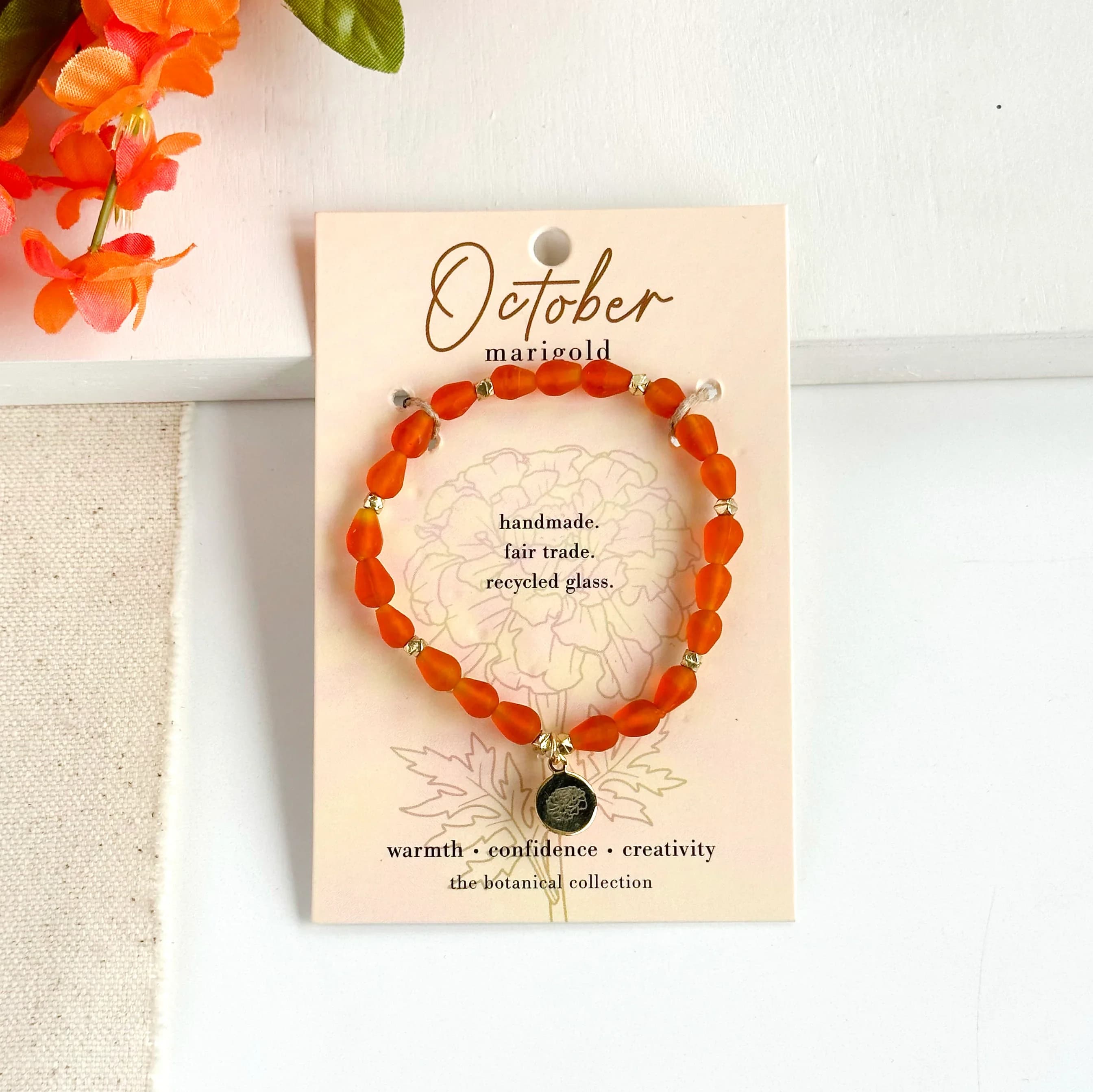 October-Birth Flower Bracelet - Hello, October!  Part of our Botanical Collection, the October Bracelet is crafted from bright orange glass beads, brass accent beads, and features a marigold charm. Great for gifting, the card features the marigold and it's symbols of warmth, confidence and creativity, along with a garden-inspired quote. This bracelet also benefits an urban community garden project!  The semi-opaque beads have a satin, frosted texture and are handmade from recycled glass. The flower charm is 24k gold plated.   Sustainably handmade by women artisans in India.   