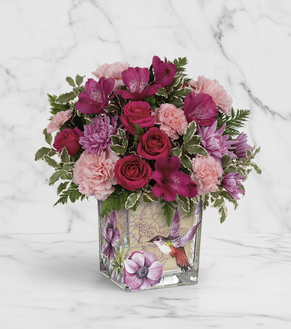 Pink and magenta flowers arranged in a square glass vase with a bird design