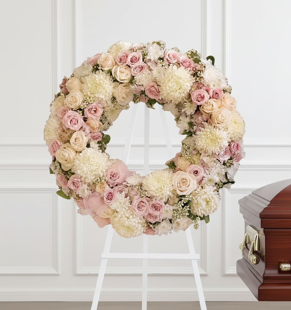 Pastel floral wreath on a white stand beside a wooden casket
