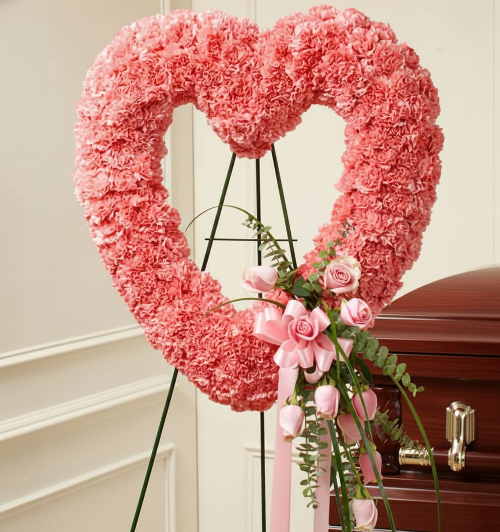 Pink heart-shaped floral wreath on an easel beside a casket