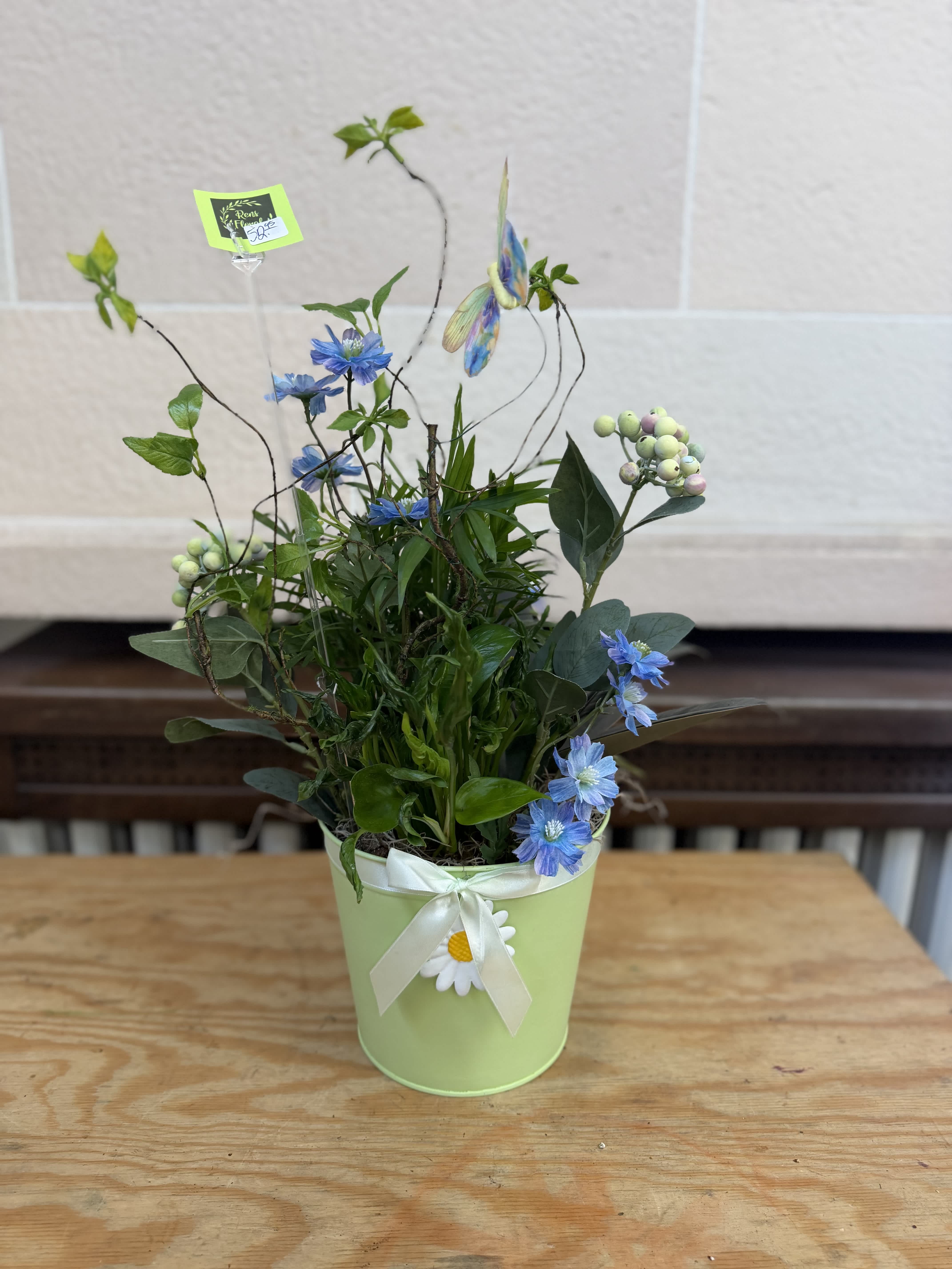 Blue flowers in a light green pot with a white ribbon and daisy accent