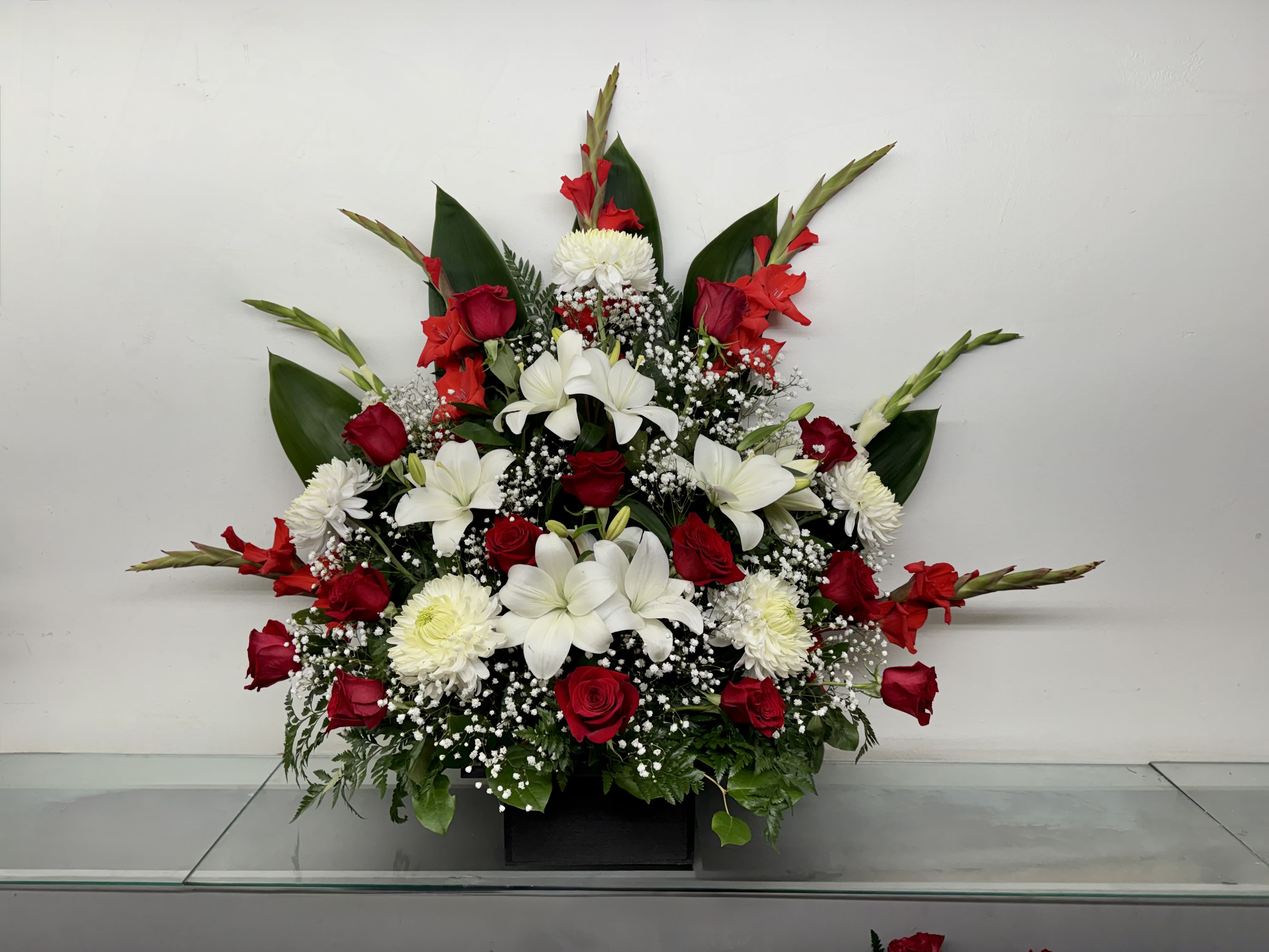 Red and White Elegance - A striking one-sided arrangement featuring radiant red roses, elegant white lilies, crisp white chrysanthemums, and vibrant red gladiolus accented with delicate baby's breath and lush greenery. Designed with height and balance, this arrangement makes a beautiful statement for podium displays, commemorative events, celebrations of life, anniversaries, and special occasions.