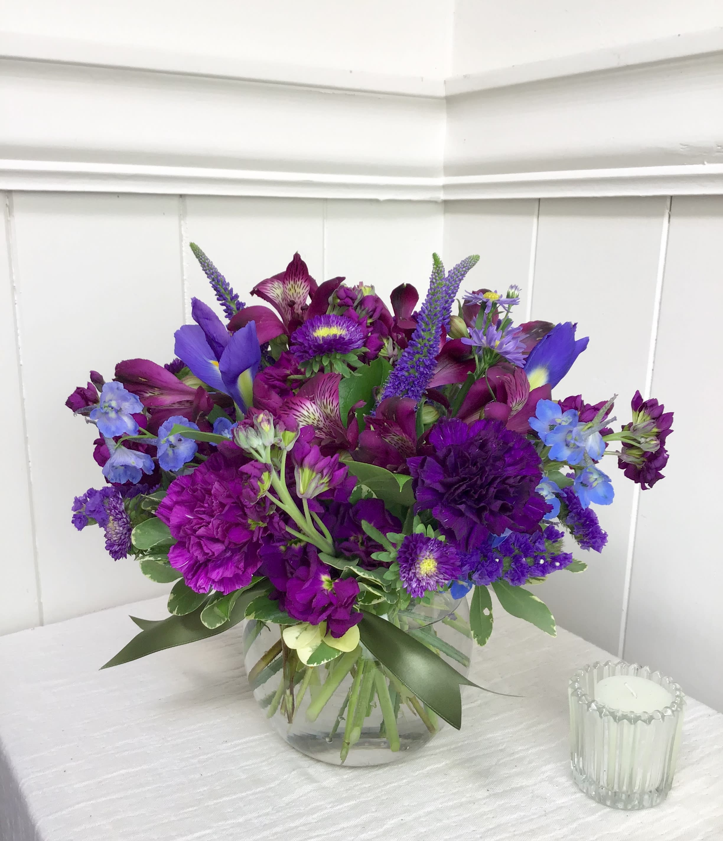 Round arrangement of purple and blue flowers in a clear glass vase on a white table.