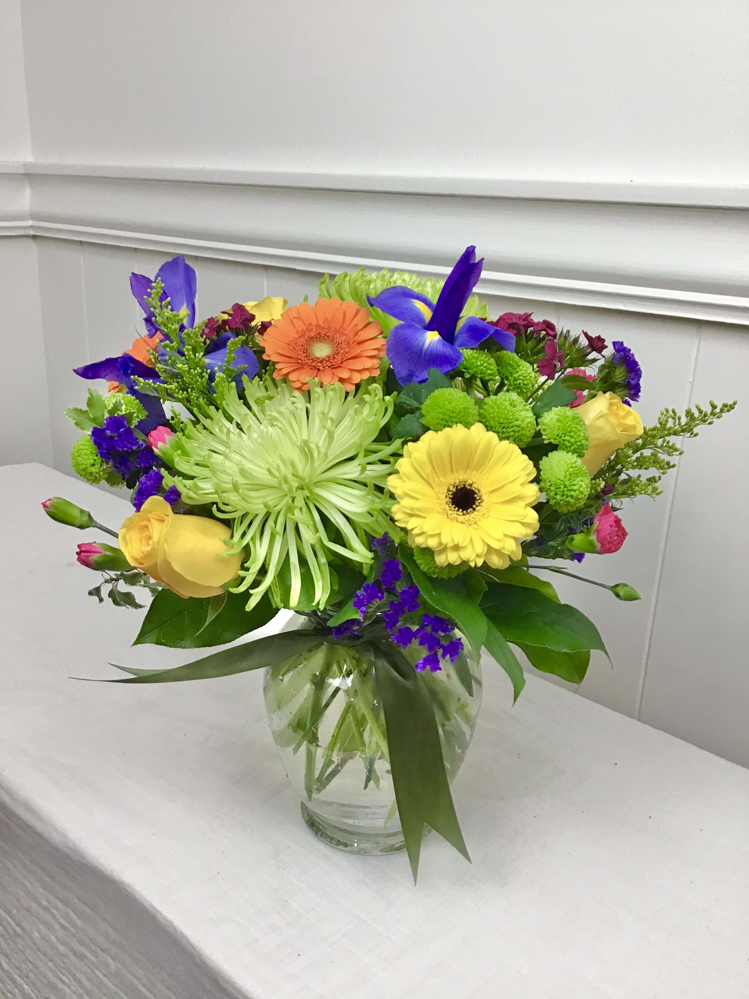 Bright mixed bouquet with yellow roses, gerbera daisies, green spider mums, and purple irises in a glass vase.