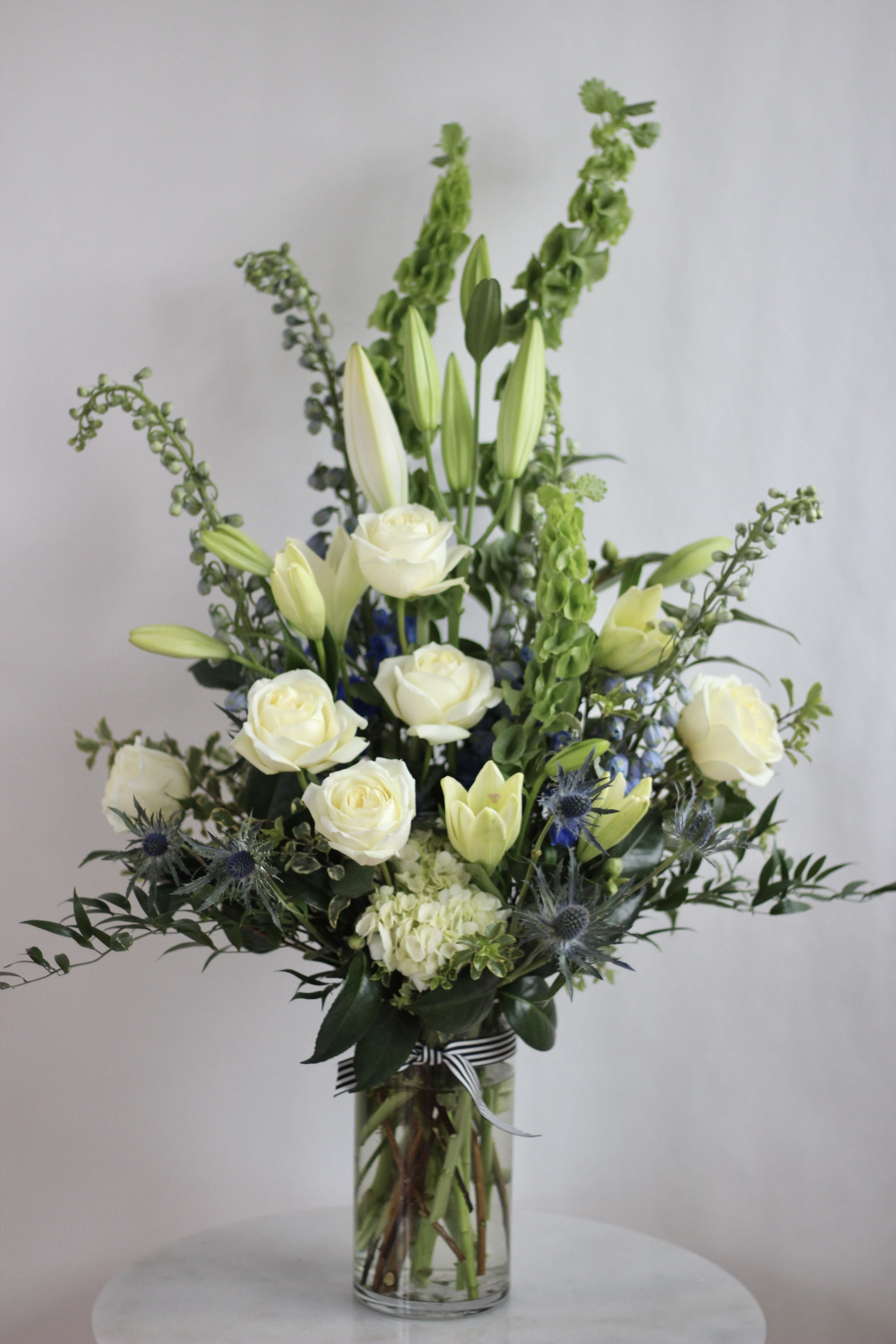 White roses and lilies in a tall glass vase with blue thistle accents