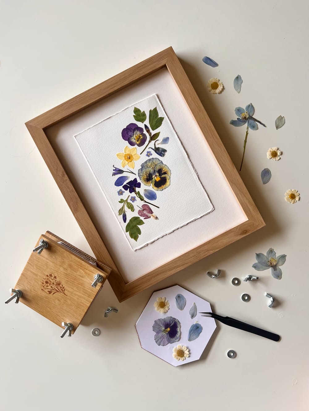 Pressed flowers arranged in a wooden frame with loose petals and tools nearby.