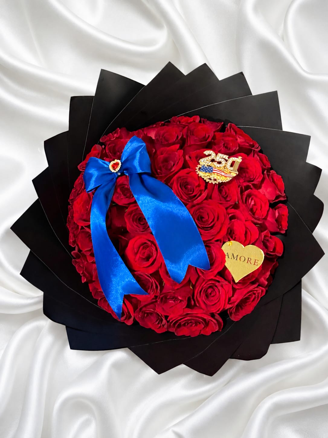 USA 250 Red Rose Heart – Limited Edition - Celebrate America’s 250th anniversary with our exclusive USA 250 Red Rose Heart – Limited Edition. This elegant arrangement features premium fresh red roses, luxurious black wrapping, and a royal blue satin bow for a sophisticated patriotic presentation.  The centerpiece of this design is a beautiful USA 250 commemorative brooch, celebrating this historic milestone. The brooch is removable and can also be worn as a stylish clothing accessory, creating a lasting keepsake long after the flowers have been enjoyed.  Handcrafted by AMORE Flowers, this luxury arrangement is perfect for Independence Day celebrations, birthdays, anniversaries, military families, patriotic events, or anyone who wants to honor America’s 250th anniversary with an unforgettable floral gift.