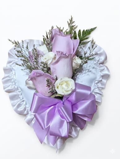 Heart Casket Pillow - A compact, handcrafted heart casket pillow designed to offer a gentle, comforting tribute for sympathy and funeral services. This flower arrangement features a soft palette of white and purple flowers. A thoughtful token to honor a loved one and convey warmth and remembrance.  The pillow is accented by lavender roses and white sweetheart roses, fine white filler, and bright green foliage for contrast. Glossy satin ribbon, a large lavender bow, and a ruffled decorative fabric wrap give the piece a formal polished presentation.  This design makes a small but personal statement at a service or wake and can be customized by color on request. 