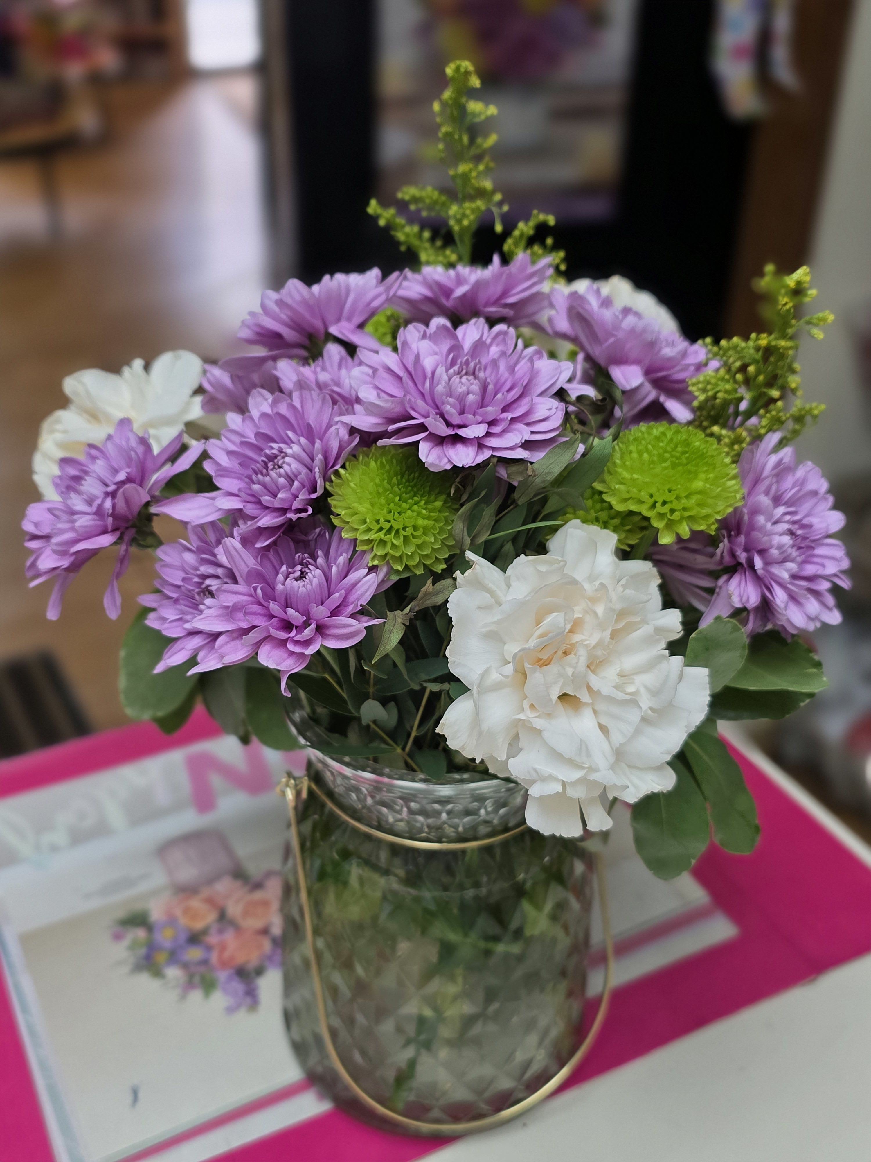 Candle of Dreams - Chrysanthemums, carnations set a cheerful, romantic tone in this flower arrangement for birthdays, anniversaries, or any sunny celebration. The mix of warm peach carnations, cool purple accents, and small pale pink carnation-like blooms creates a lively yet balanced palette that will make the person you're thinking of feel genuinely celebrated.  Loosely gathered in a clear glass vase and finished with a soft color ribbon, the bouquet has a fresh, hand-tied look with airy greenery that adds movement and texture. Includes roses, chrysanthemums, carnations and complementary accent blooms in pink, purple, white and green-an elegant, versatile choice for sending warm wishes or brightening someone's day. 